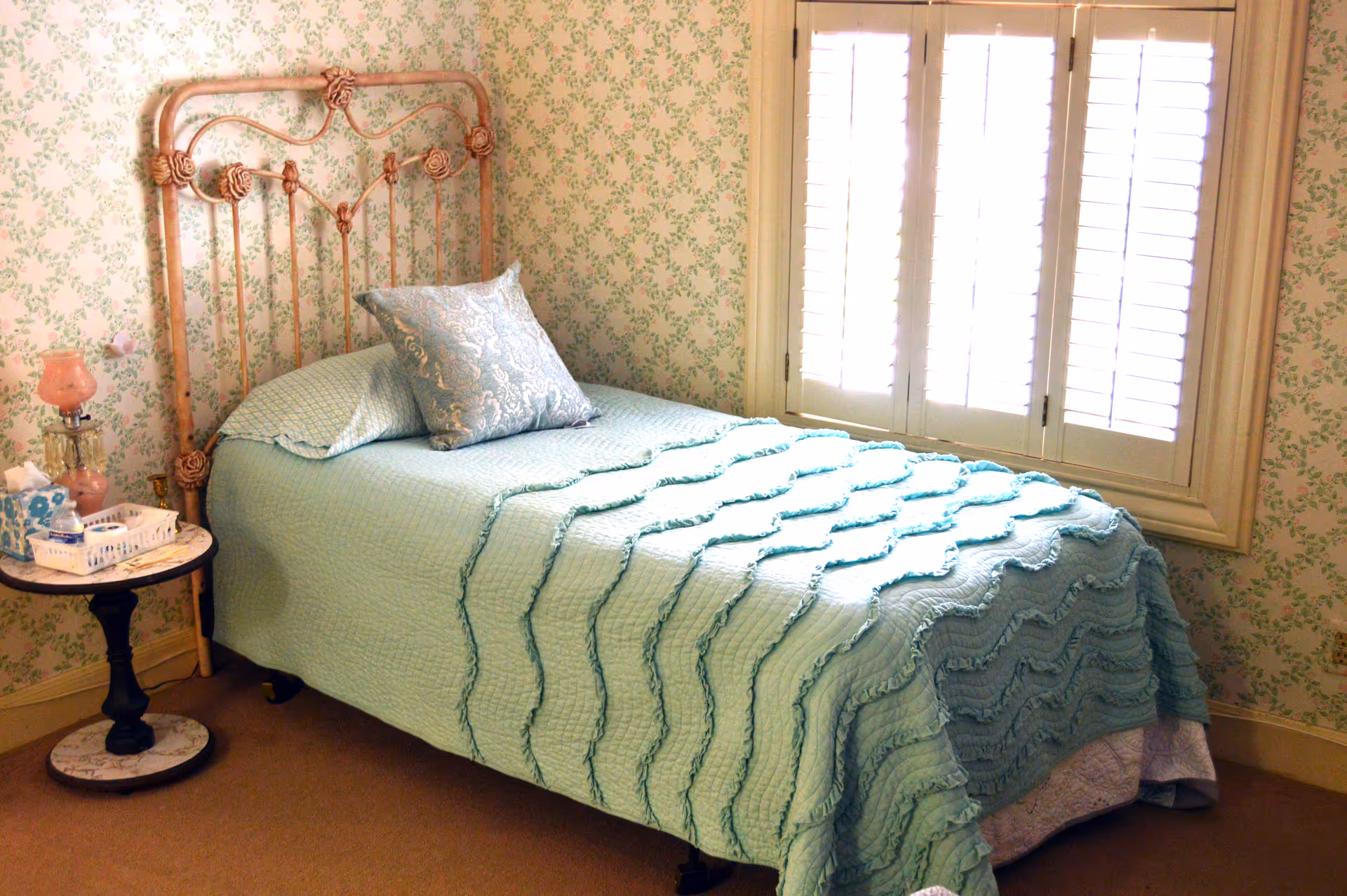 Pastel bedroom with teal quilted bed, ornate metal headboard, and sunlit window blinds.