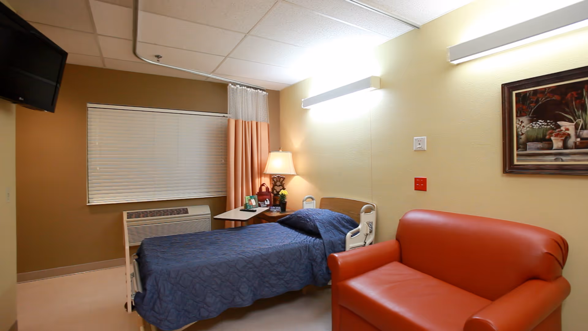 A small, neatly arranged bedroom in a care facility featuring a single hospital-style bed with a blue quilt, a red leather loveseat, a bedside table with a lamp and framed photos, a window with closed blinds and peach-colored curtains, and a wall-mounted TV in the corner.
