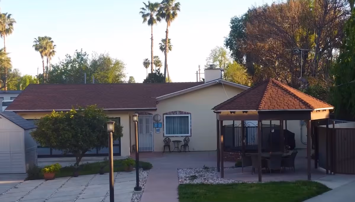 Single-story building with a brown roof and light-colored walls, featuring a small patio area with a table and two chairs. In front of the building, there is a paved walkway flanked by two lamp posts, a small tree, potted plants, and a gazebo with a red-tiled roof and outdoor seating underneath. Tall palm trees and other greenery are visible in the background under a clear sky.
