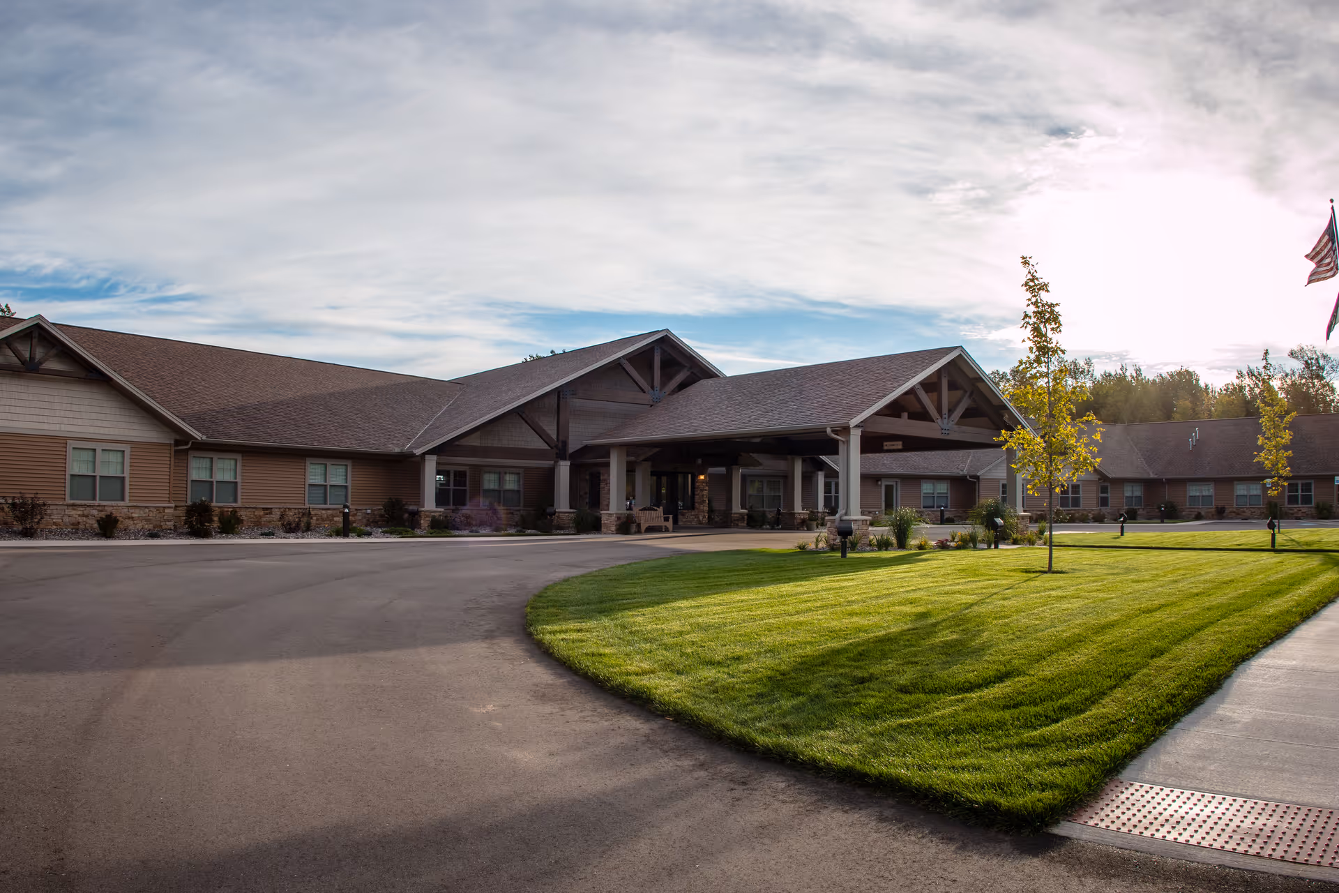 Exterior view of Teal Lake Senior Living Community building with a large covered entrance, well-maintained green lawn, young trees, and a partly cloudy sky.
