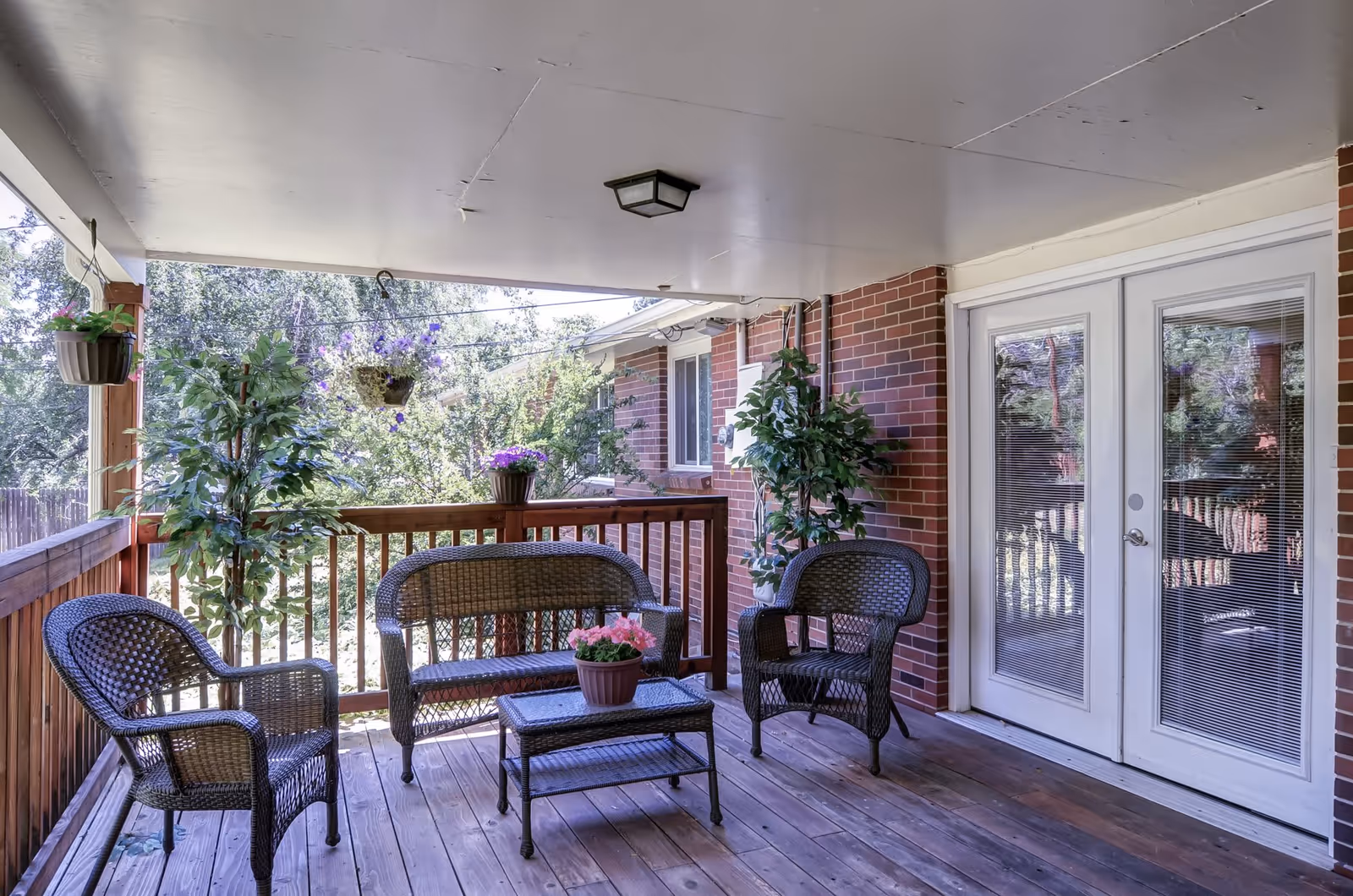 Covered outdoor patio area with wooden floor and railing, featuring two wicker armchairs, a wicker loveseat, and a wicker coffee table with a potted pink flower. There are hanging flower pots and potted plants around the patio. The patio is attached to a brick building with white double doors having glass panels with blinds.