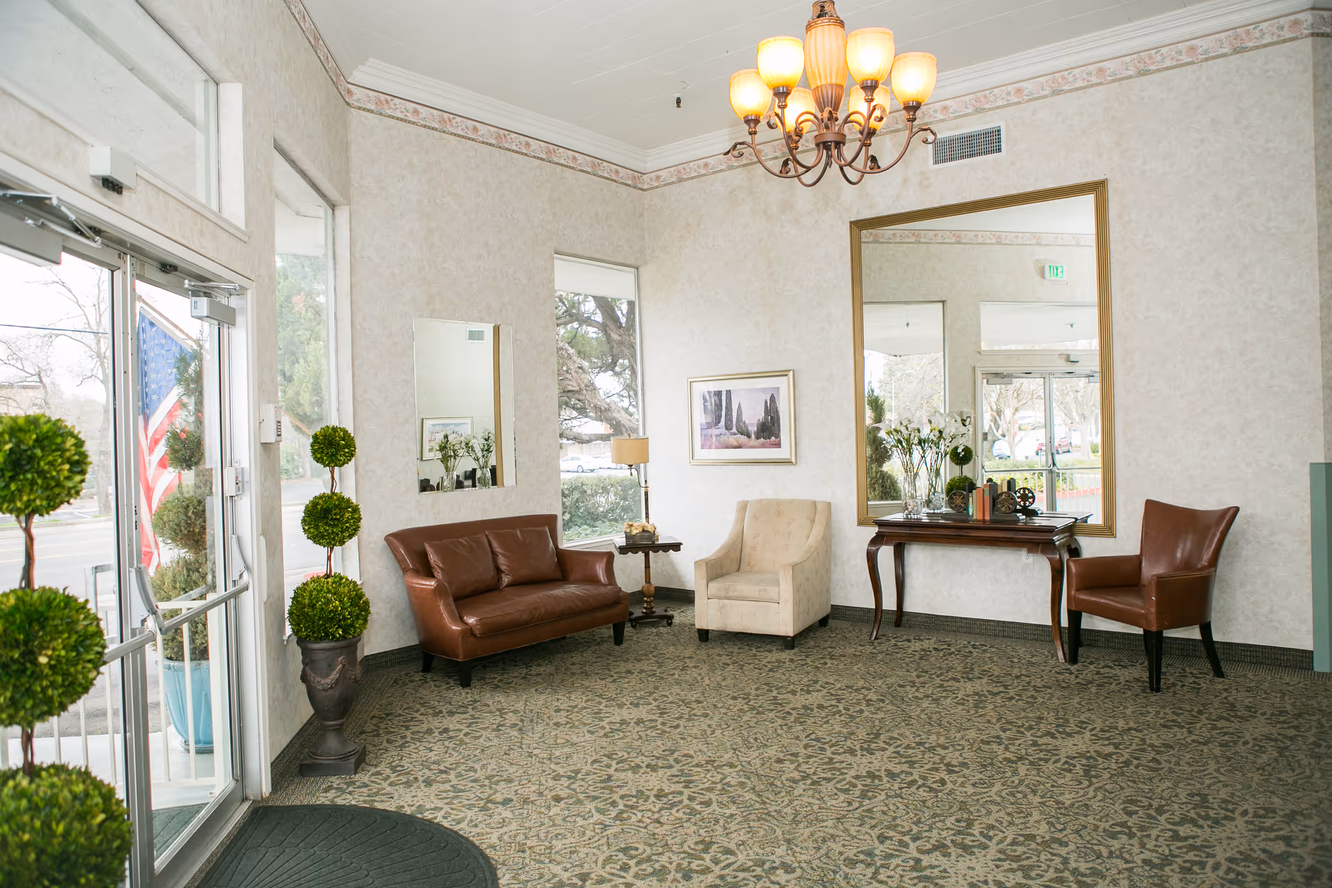 A bright and welcoming lobby area with a glass entrance door on the left, decorated with potted topiary plants. Inside, there is a brown leather loveseat, a beige armchair, and a brown leather chair arranged around a small wooden table with a lamp. A large mirror hangs on the wall above a wooden console table adorned with decorative items and flowers. The room has patterned carpet, light-colored wallpaper, and a chandelier hanging from the ceiling.