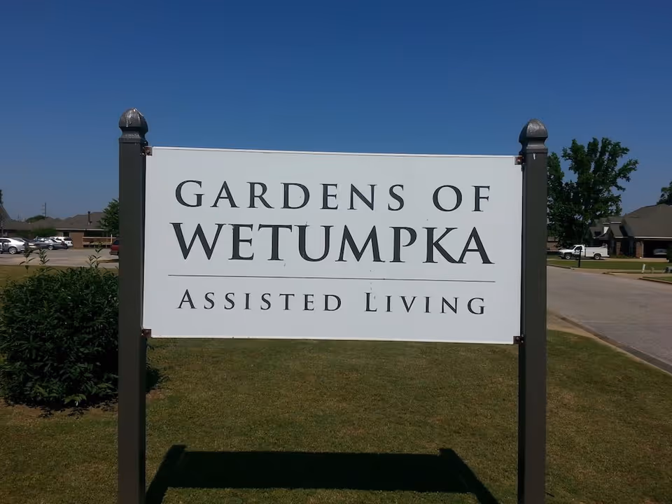 Large white sign reading Gardens Of Wetumpka Assisted Living in a grassy front yard.