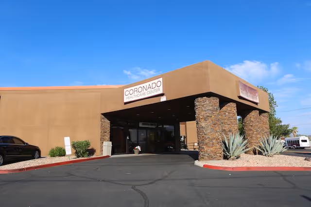 Exterior front view of Coronado Healthcare Center building with a covered entrance supported by stone pillars, desert landscaping with agave plants, and a clear blue sky.