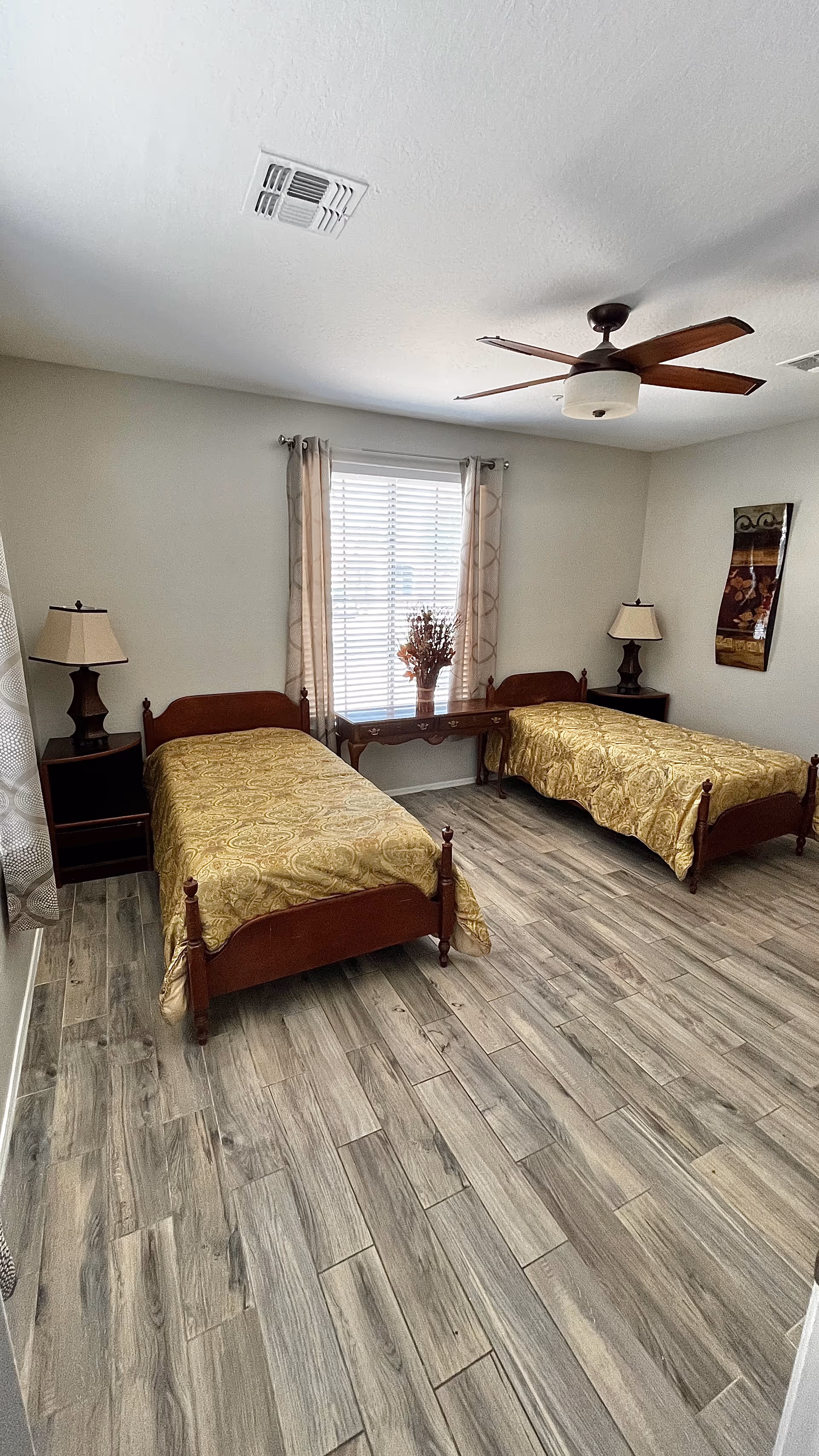 Two single beds with matching gold bedspreads flank a desk beneath a window and ceiling fan.