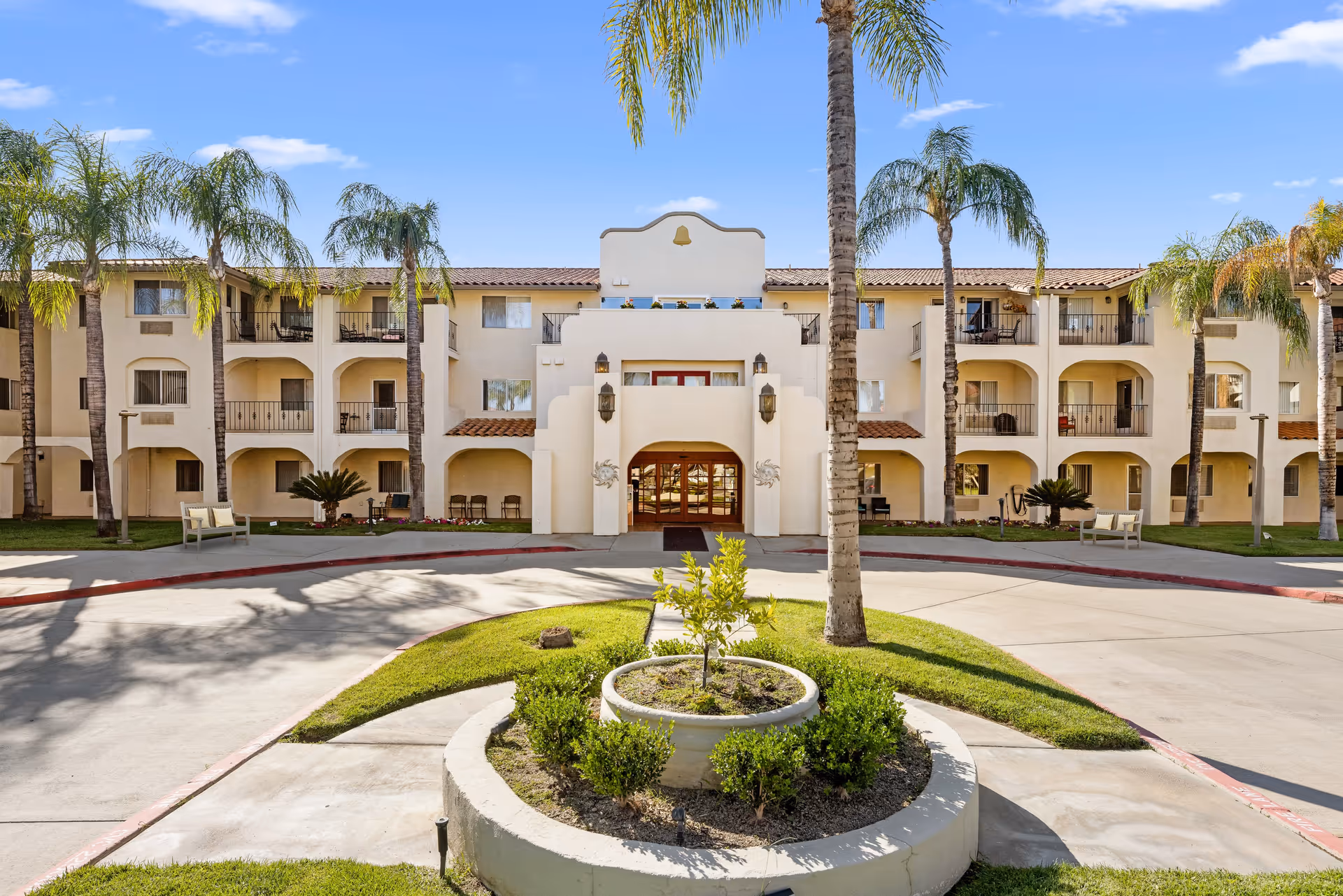 Front exterior view of Mission Commons - A Provincial Senior Living Community, a three-story building with balconies, palm trees, and a circular driveway with landscaped greenery in the center.