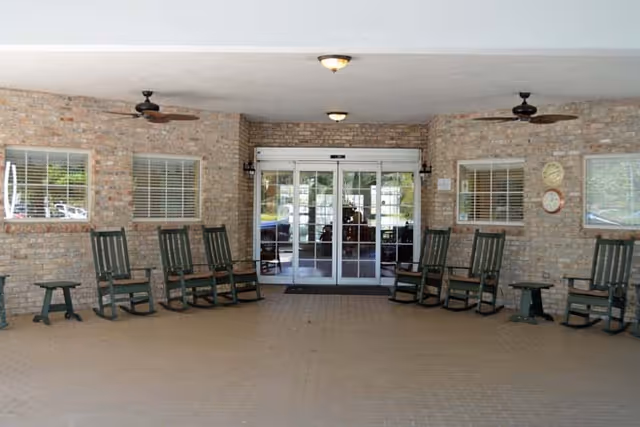 Covered entrance area with brick walls and a tiled floor, featuring several green rocking chairs and small tables arranged along the walls. Two ceiling fans and ceiling lights are mounted overhead. Glass automatic sliding doors lead inside the building.