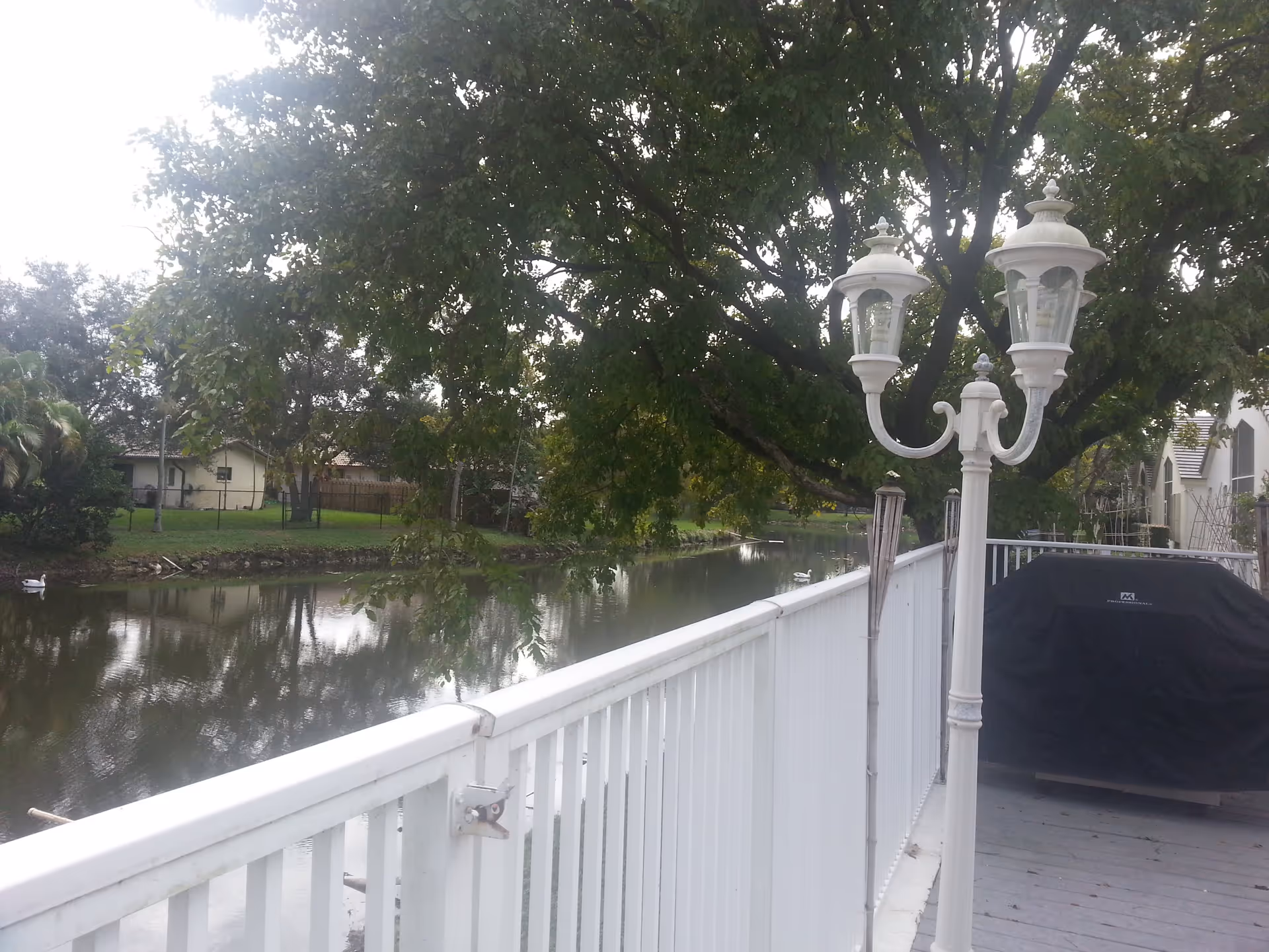 White deck with railing and vintage-style lamp posts overlooking a tree-lined canal and houses, with a covered grill on the right.