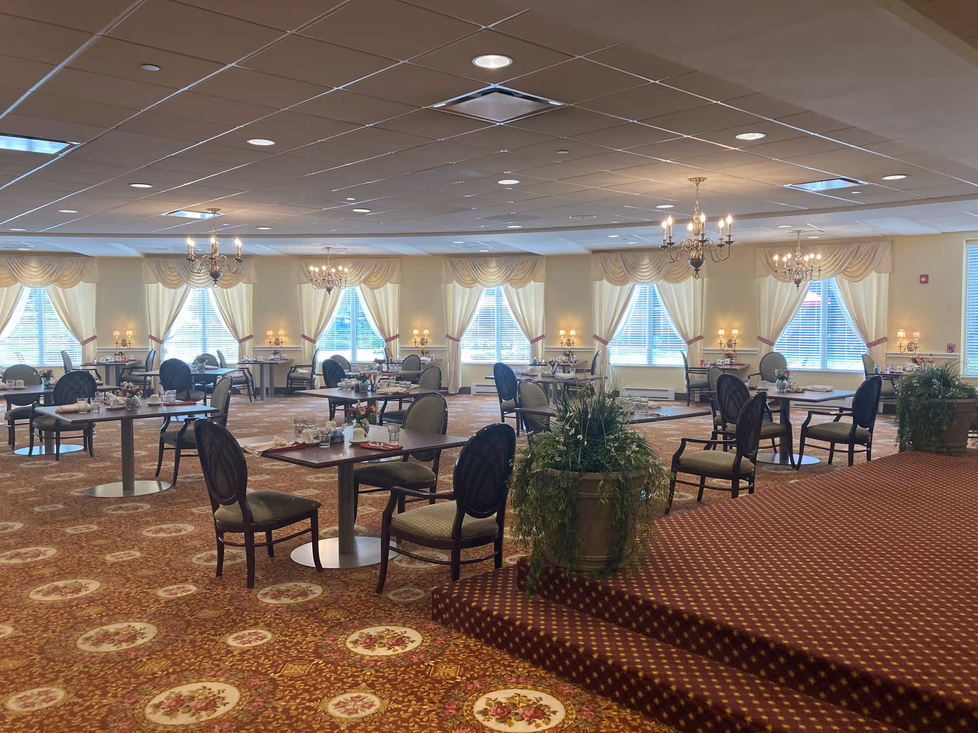 A spacious dining room with multiple round tables and chairs arranged neatly. The room features large windows with cream-colored curtains, chandeliers hanging from the ceiling, and floral-patterned carpet. There are decorative plants and table settings with glasses and napkins on each table.