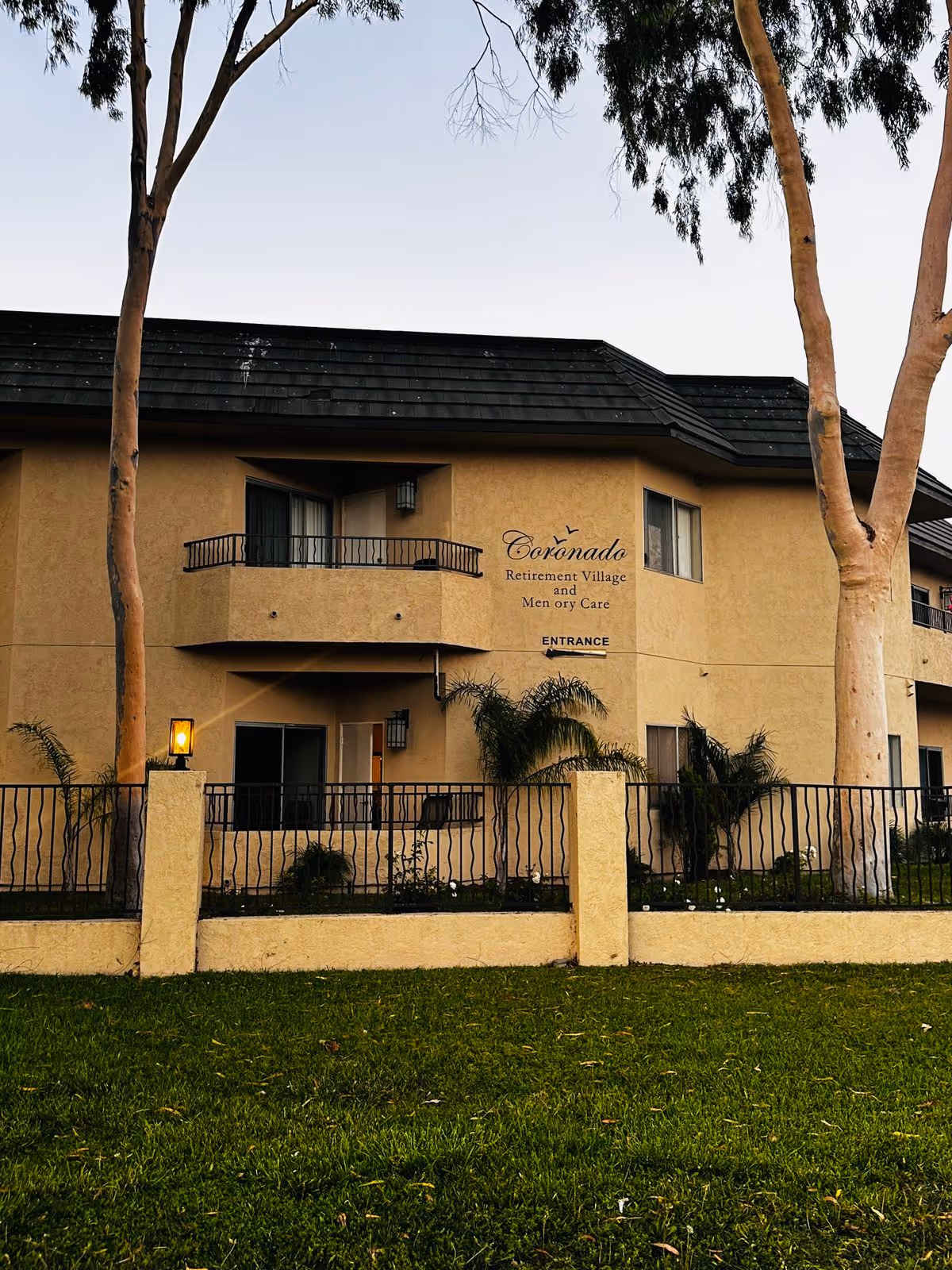 Front exterior of a two-story beige building with balconies and a sign reading 'Coronado Retirement Village' behind a fence and lawn.