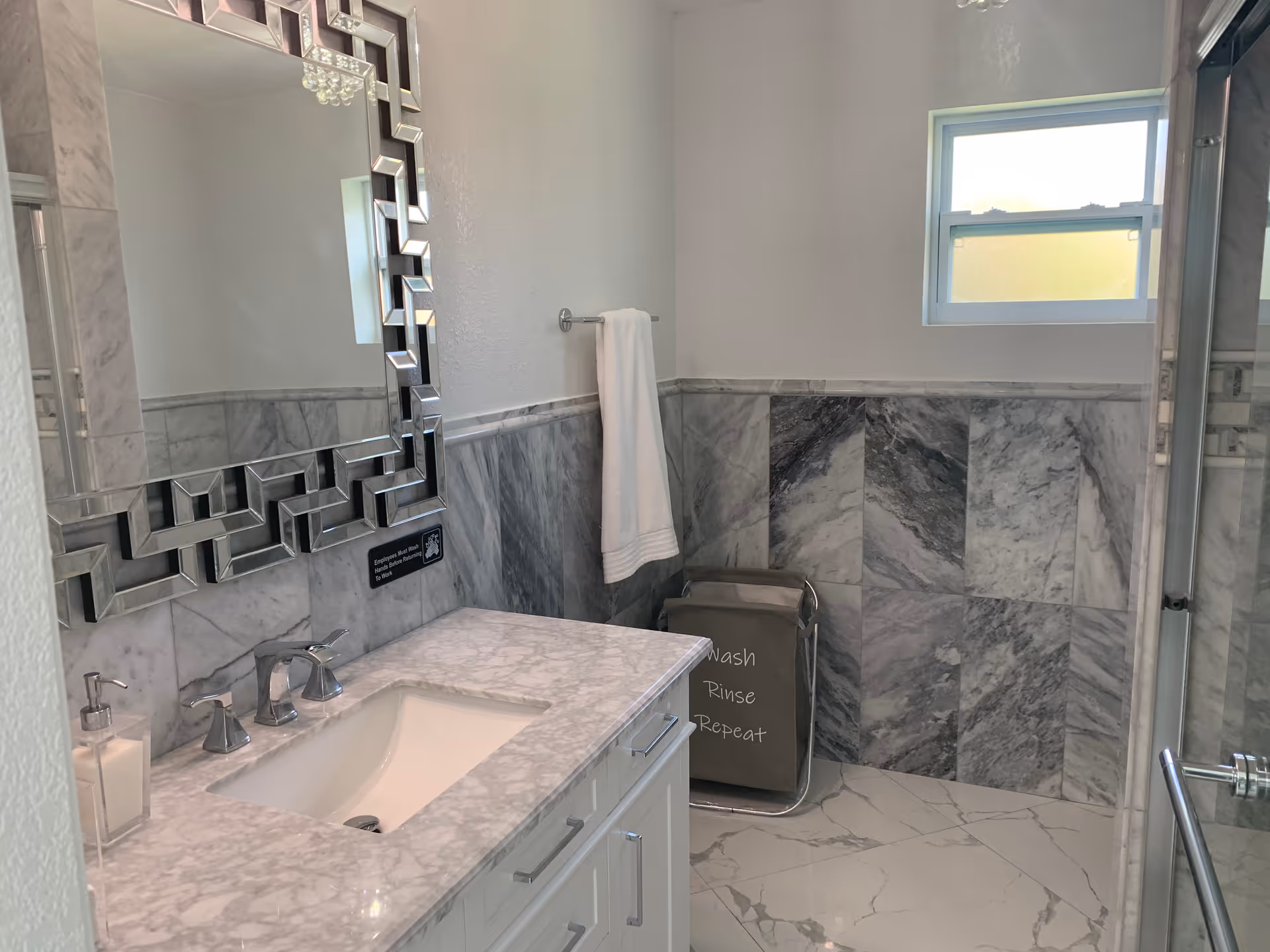 Marble tiled bathroom with dual sink, geometric mirror, towel bar, window, shower, and laundry hamper.