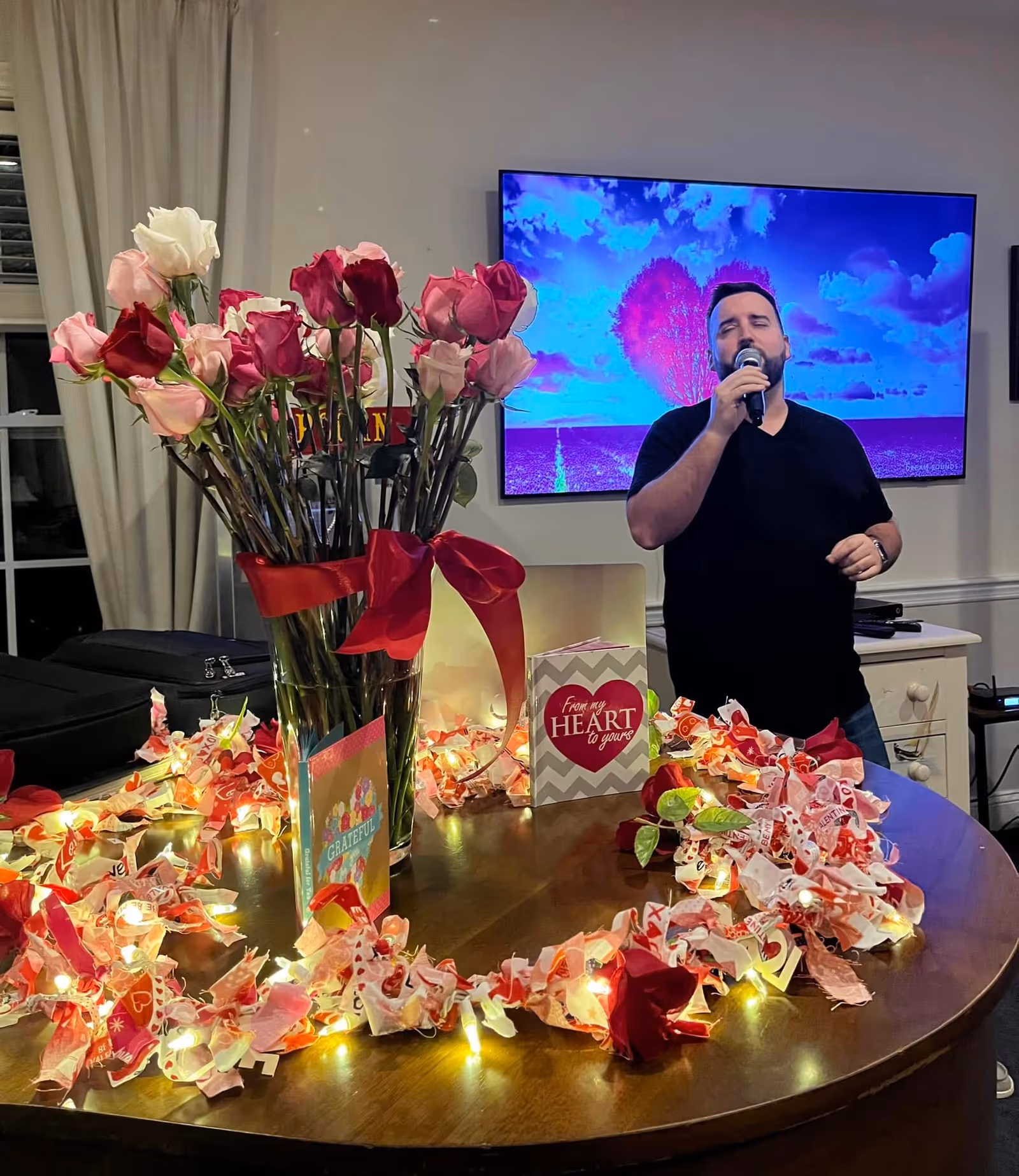 A man singing into a microphone in a room decorated with a vase of pink and red roses, Valentine's Day themed cards, and a string of lights with red and white fabric on a wooden table. A large TV screen in the background displays a colorful image of a tree with a heart-shaped canopy against a blue sky.