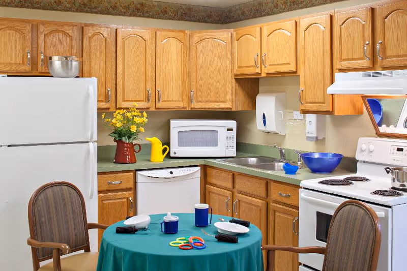 A kitchen area with wooden cabinets, a white refrigerator, microwave, dishwasher, and stove. A round table covered with a green tablecloth is set with plates, cups, and colorful rings, surrounded by two chairs. A vase with yellow flowers and a yellow watering can are on the countertop.
