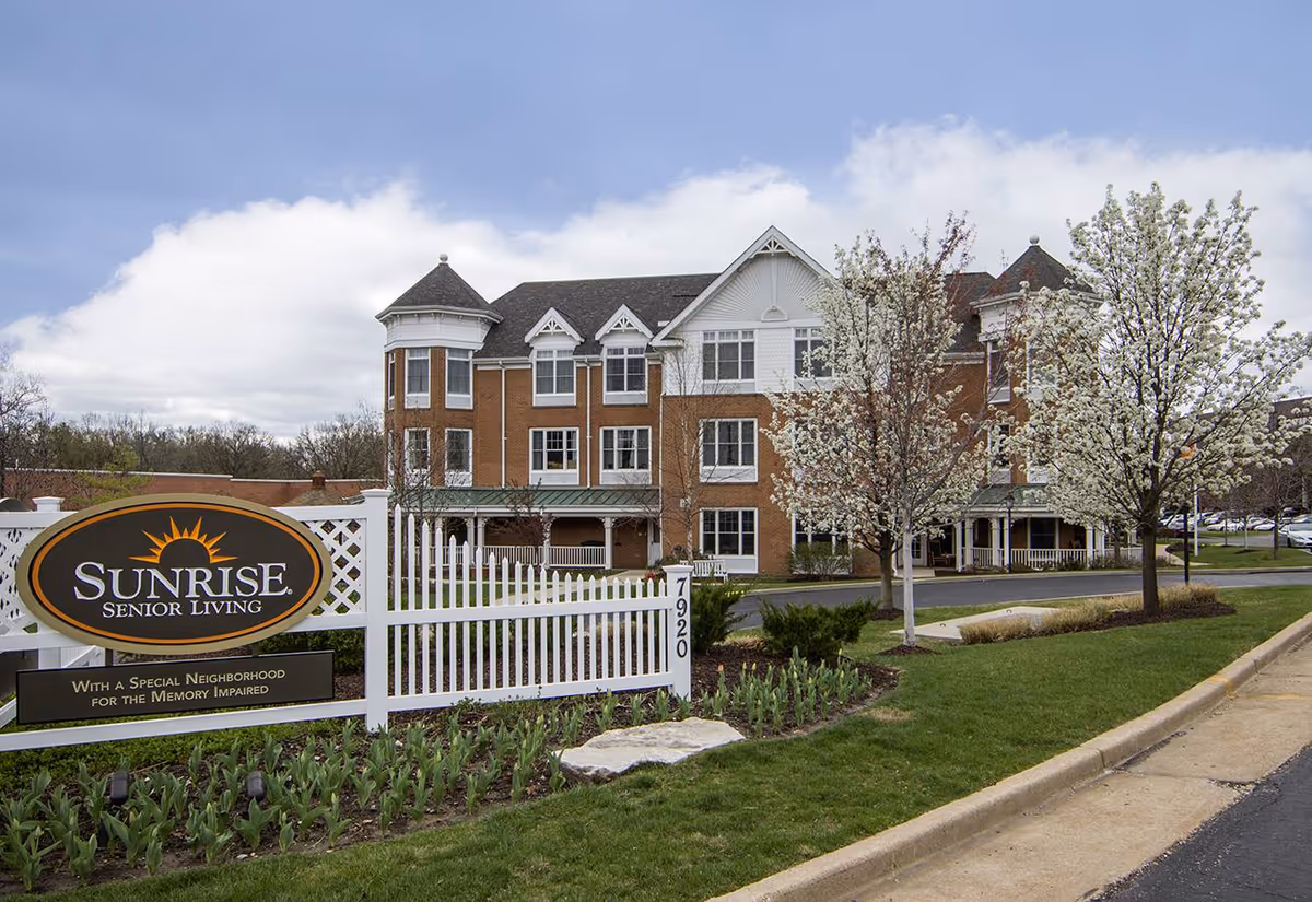 Exterior view of a multi-story senior living facility with a brick and white facade, surrounded by blooming trees and landscaped greenery. A white fence with a sign reading 'Sunrise Senior Living - With a Special Neighborhood for the Memory Impaired' is visible in the foreground.