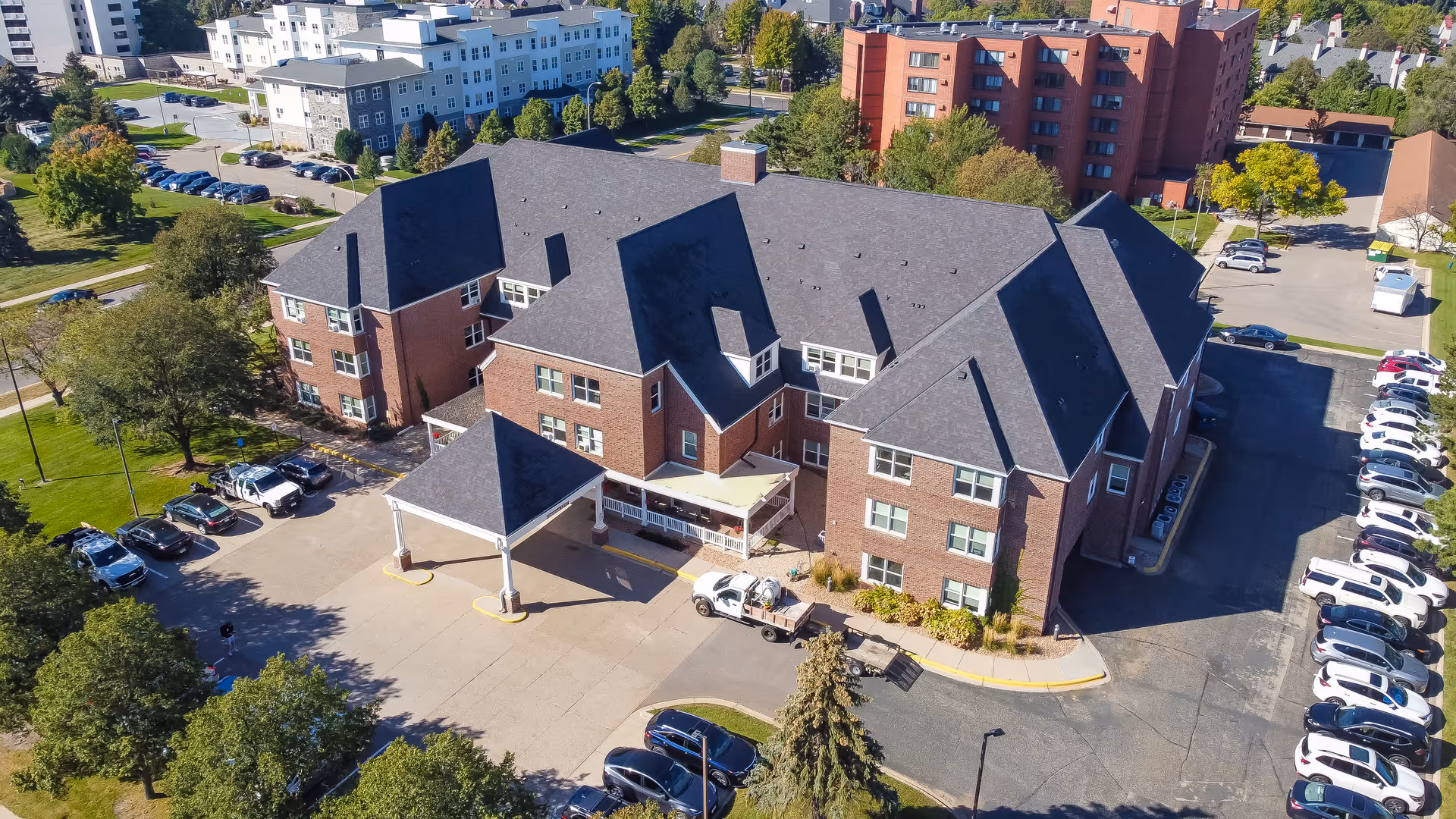 Aerial view of a brick senior living building with a covered drop-off entrance, surrounding parking lot, and nearby trees.
