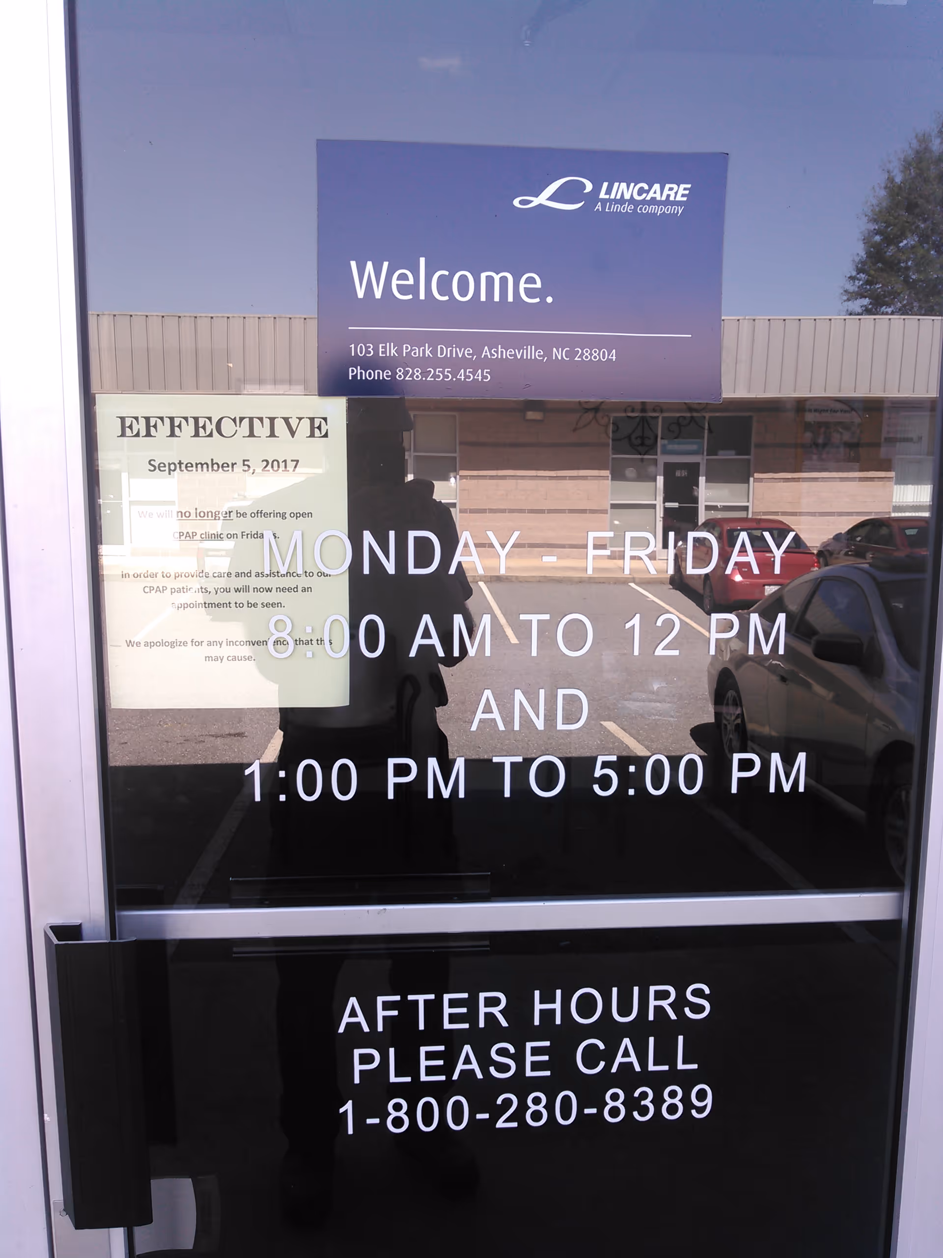 Glass entry door with Welcome sign and hours, reflecting a person and parked cars outside.