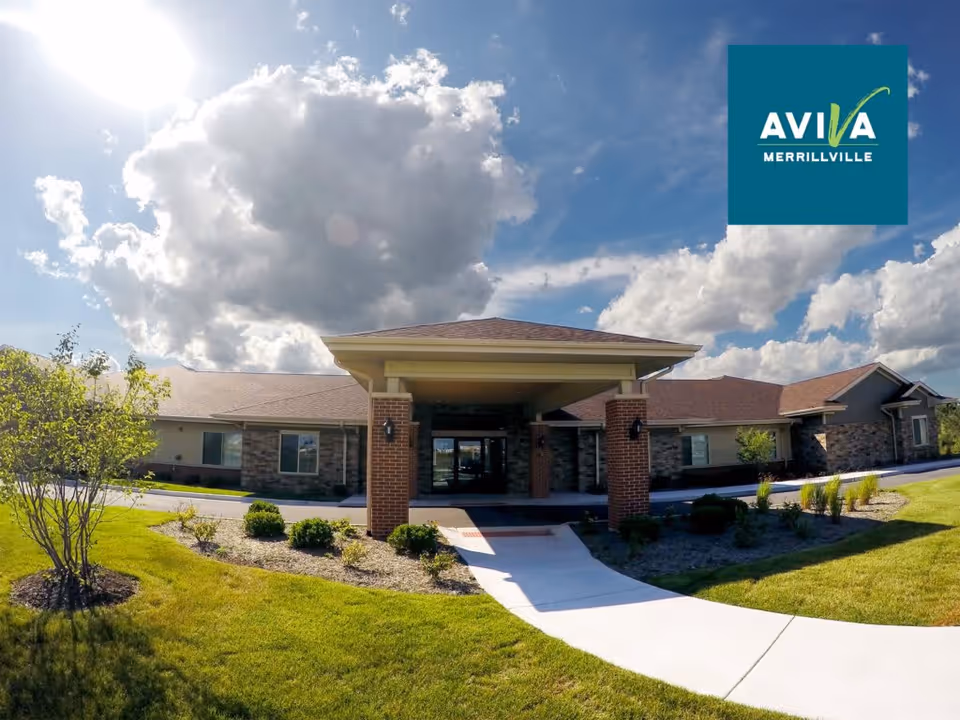 Front entrance of the AVIVA Merrillville senior living building with a covered driveway and landscaped lawn under a partly cloudy sky.