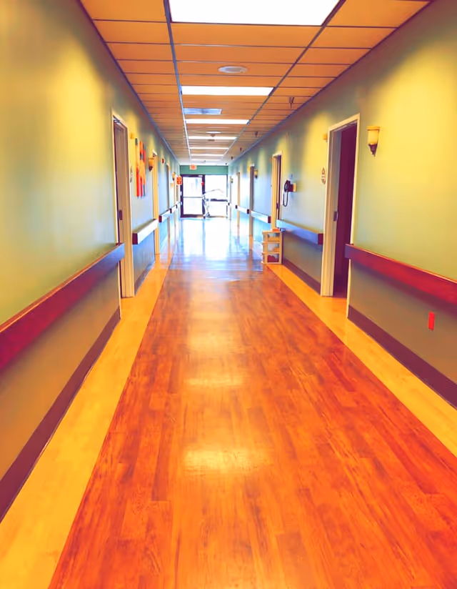 Well-lit long interior hallway in a care facility with wooden floors, wall handrails, and doors on both sides leading to rooms.