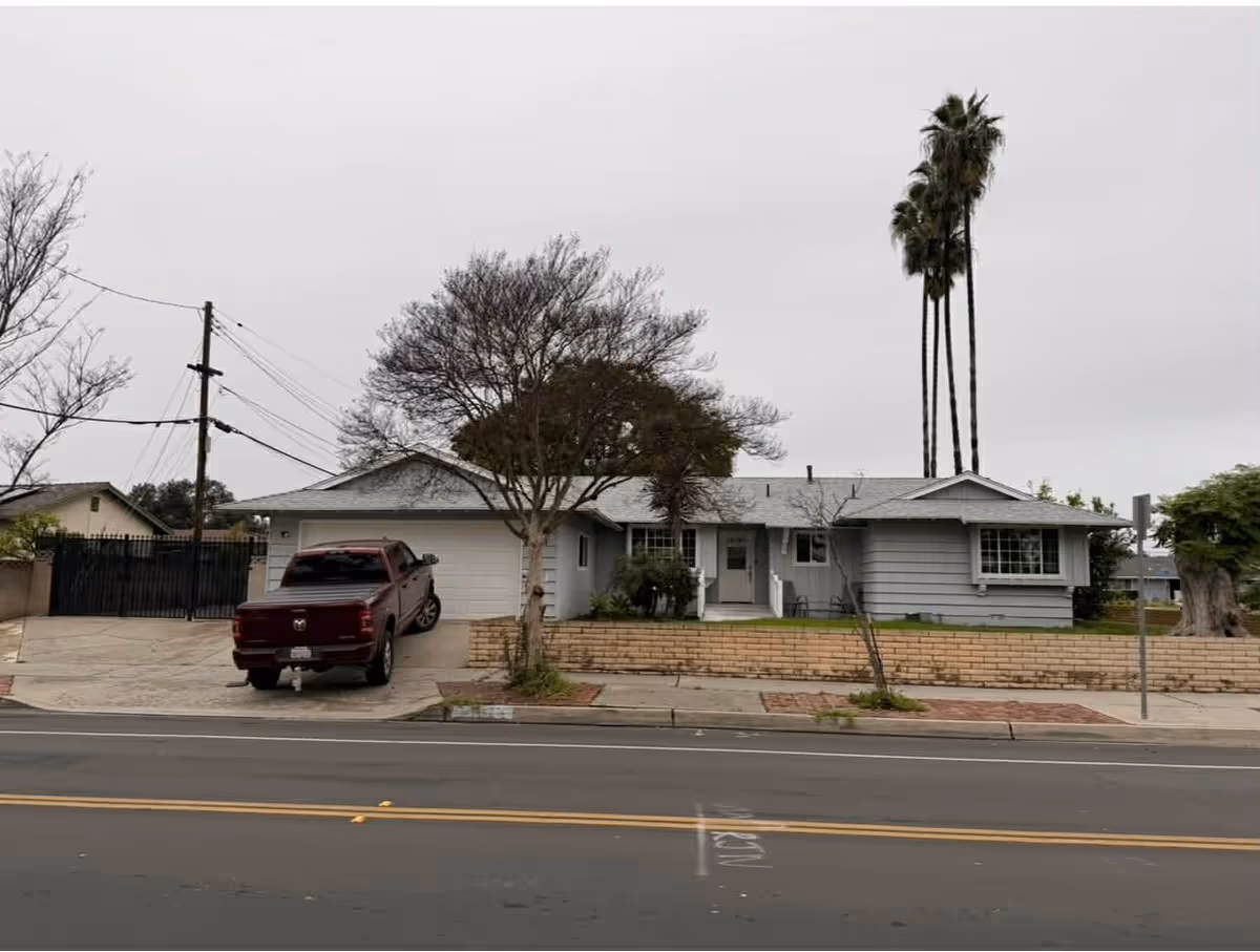 A single-story gray building with a driveway, parked truck, and tall palm trees.