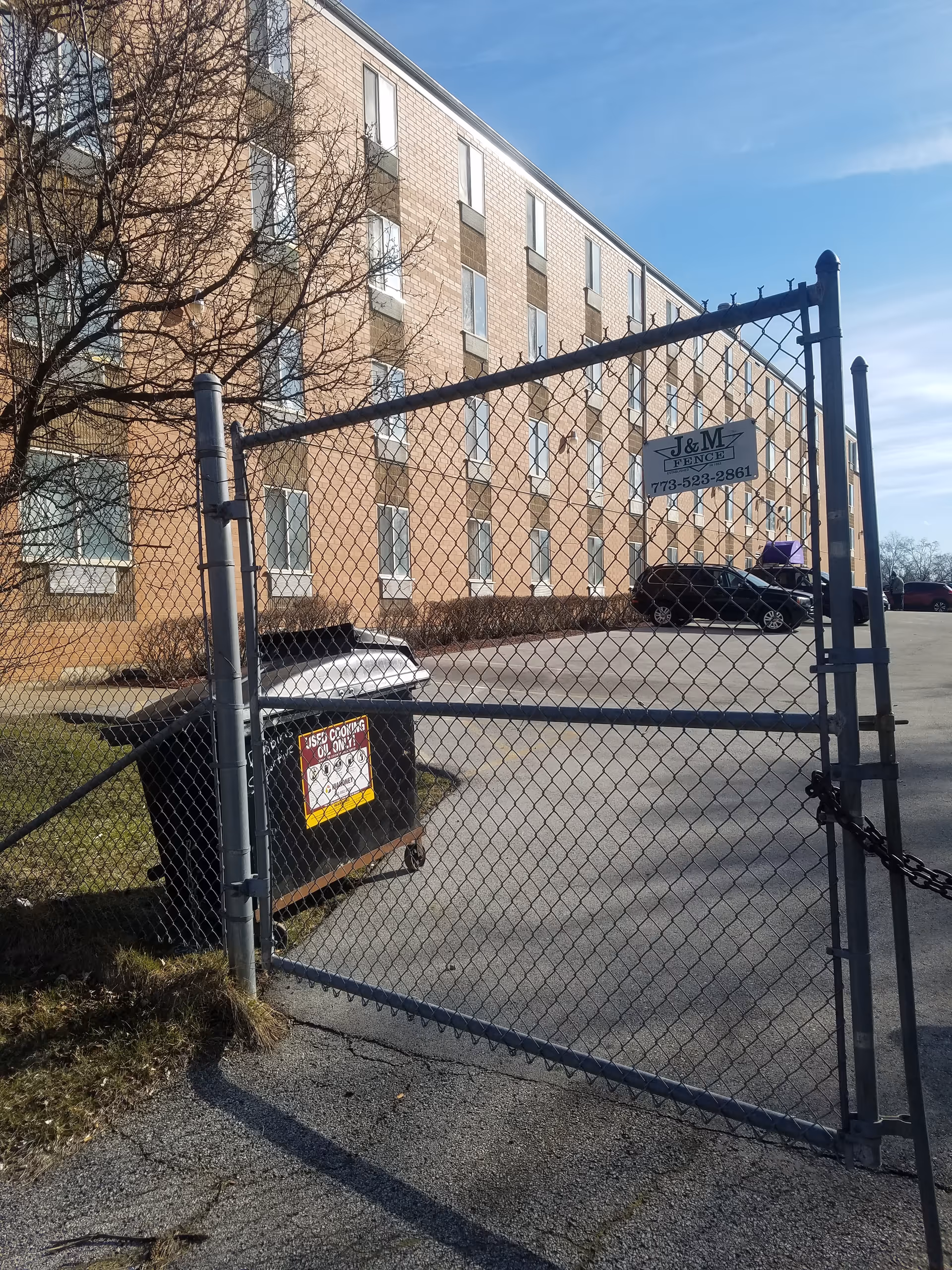 Chain-link gate in front of a brick senior living building with a dumpster nearby.