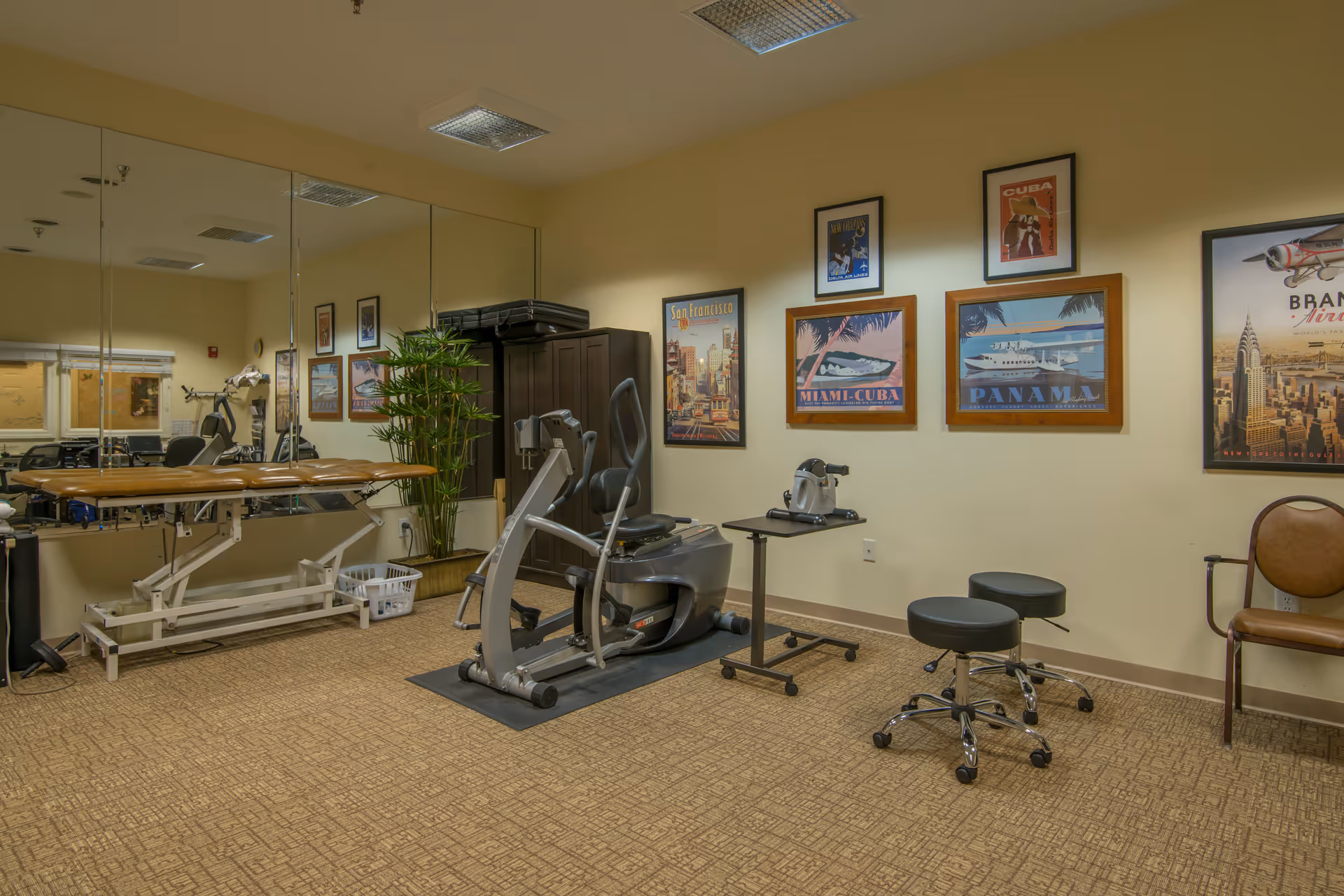 A therapy or exercise room with a recumbent exercise bike, a treatment table, two black rolling stools, a small table with exercise equipment, a large mirror on one wall, a potted plant, and framed travel posters on the walls.