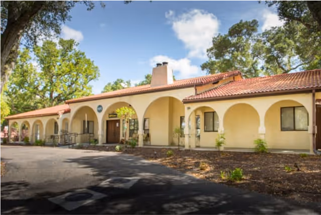 Single-story stucco building with an arched covered walkway and red tile roof set among trees under a blue sky.