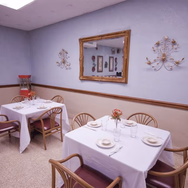 A dining room with two tables set for meals, white tablecloths and floral centerpieces.