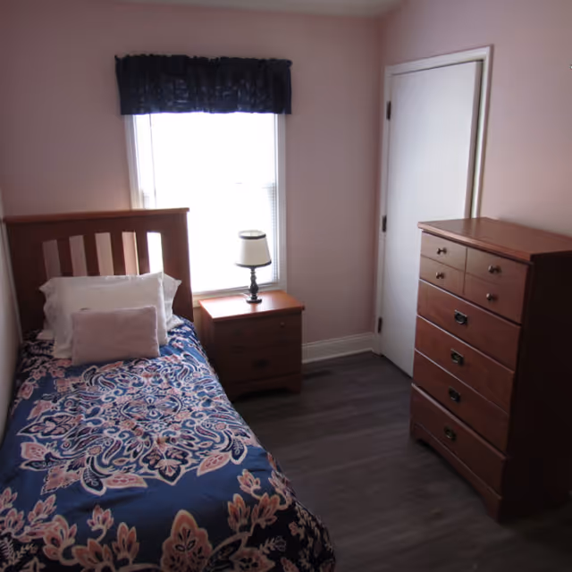 A small bedroom with a single bed covered in a blue and pink patterned bedspread, a wooden headboard, a white pillow, and a pink cushion. Next to the bed is a wooden nightstand with a lamp on it, positioned in front of a window with a dark valance. Across from the bed is a tall wooden dresser. The walls are light pink and the floor is dark wood.