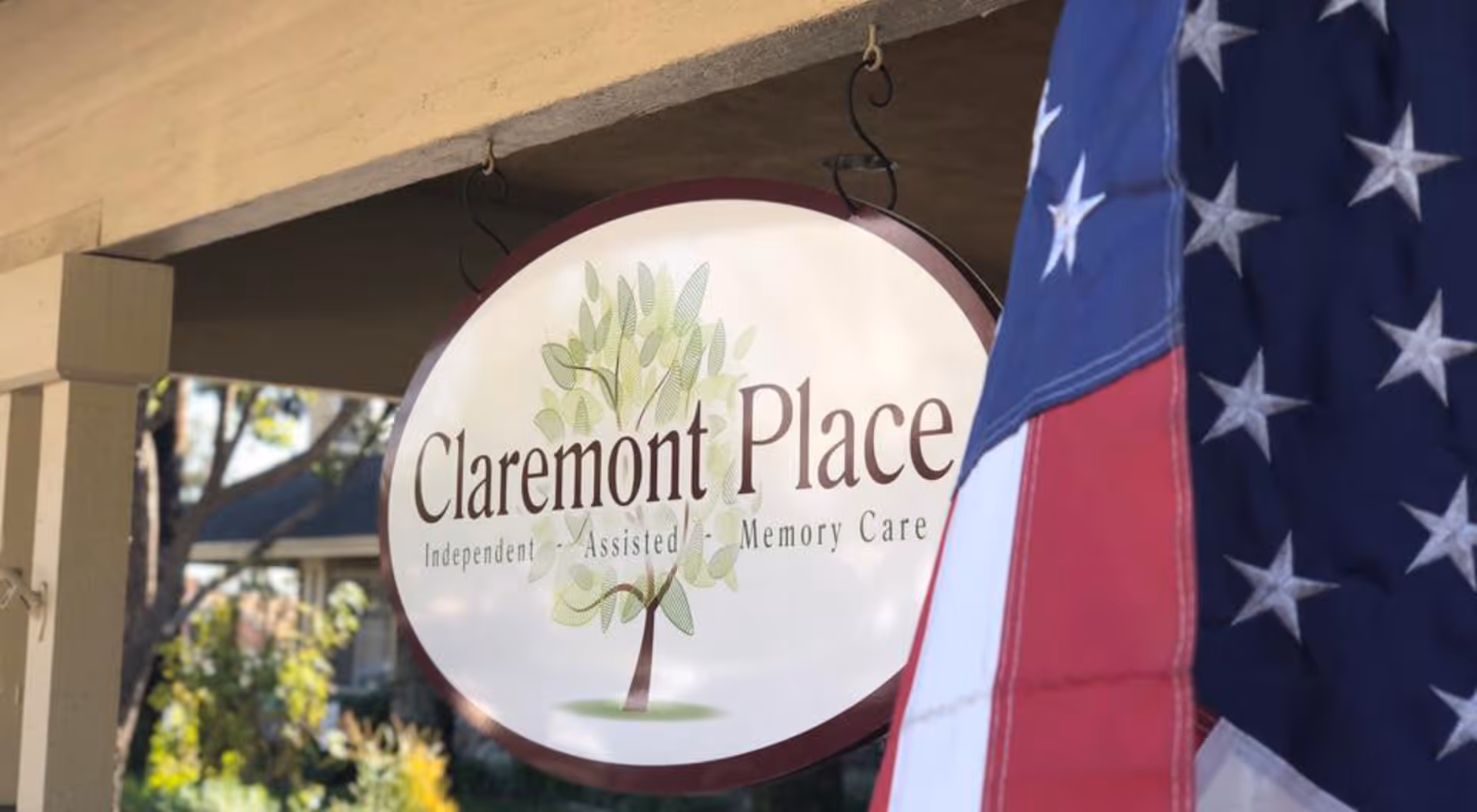A hanging oval sign for Claremont Place with a tree logo and the words Independent, Assisted, Memory Care underneath. An American flag is partially visible on the right side of the image.