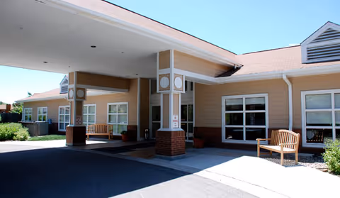 Front entrance of a single-story senior living building with a covered porte-cochere, benches, and large windows.