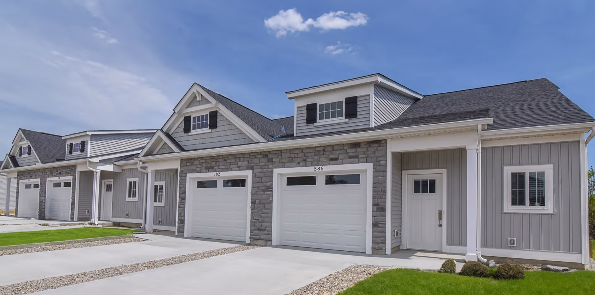 Exterior view of a modern residential building with multiple attached units, each featuring a garage with white doors, gray siding, stone accents, and small windows. The building is set against a blue sky with some clouds and has neatly maintained green lawns and concrete driveways.
