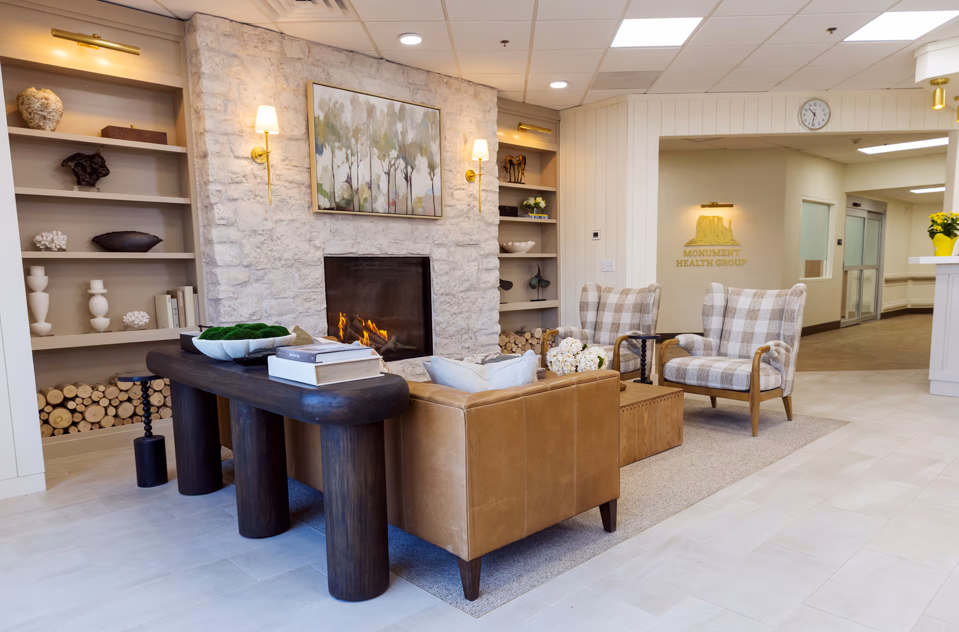 A cozy and modern living room area in Monument Health Group Millcreek featuring a stone fireplace with a fire burning, a brown leather sofa, two plaid armchairs, a dark wooden table with books and decorative items, built-in shelves with various decor, and a light-colored tiled floor.