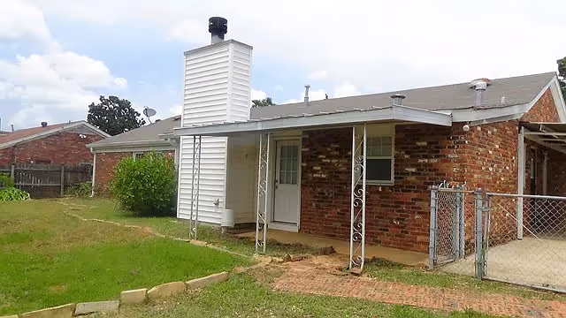 Exterior view of a single-story brick house with a small covered porch supported by decorative metal posts. The house has a white chimney structure and a fenced yard with a grassy lawn and a paved walkway.