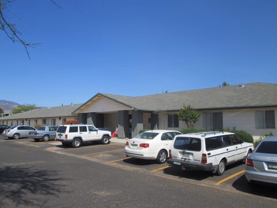 Single-story senior living building front with a parking lot and several parked cars under a clear blue sky.