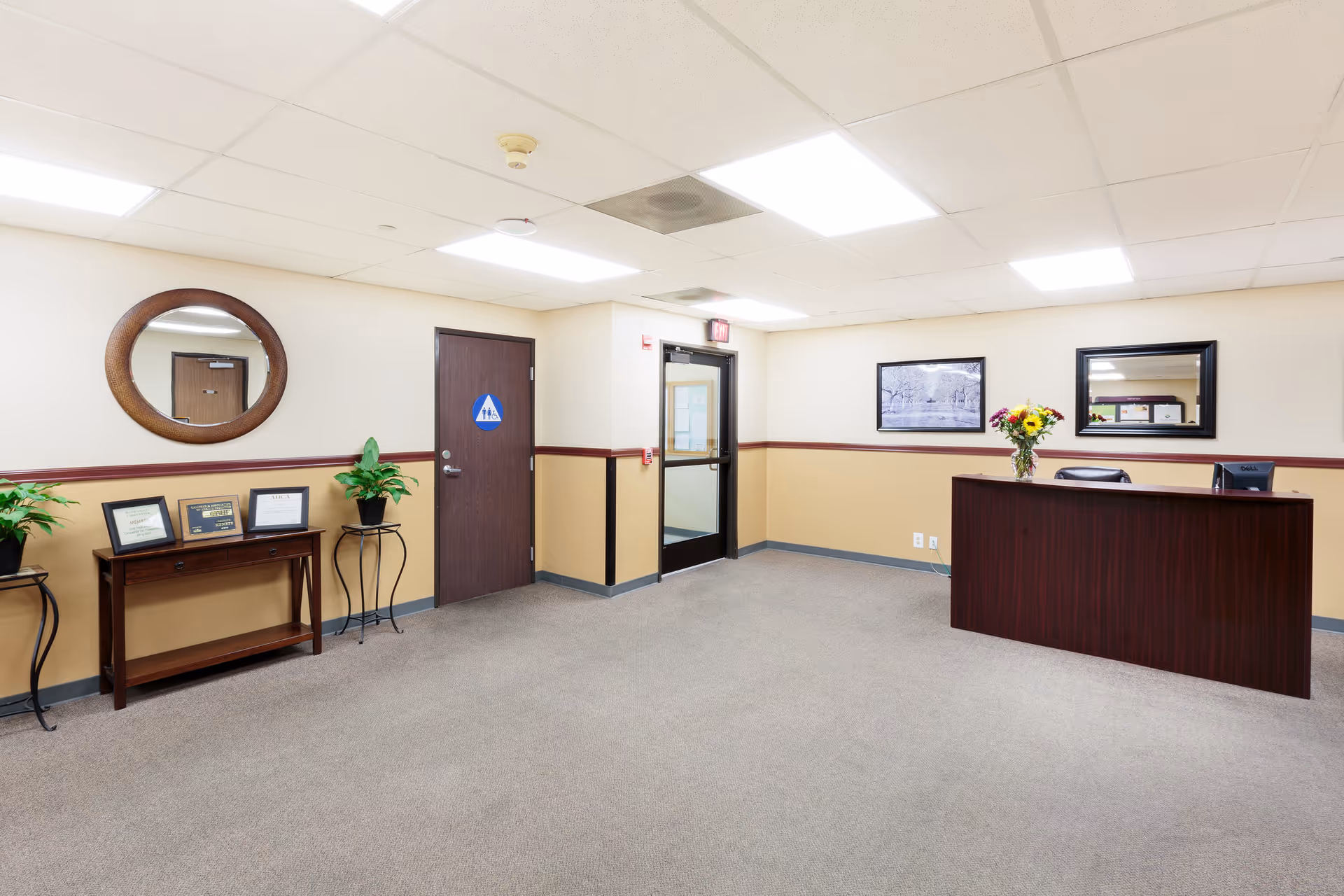 Reception area of River Valley Care Center featuring a wooden reception desk with a computer and a vase of flowers, a door with a restroom sign, a glass exit door, a round wall mirror, framed certificates on a small wooden table, two potted plants on stands, and framed artwork on the walls.