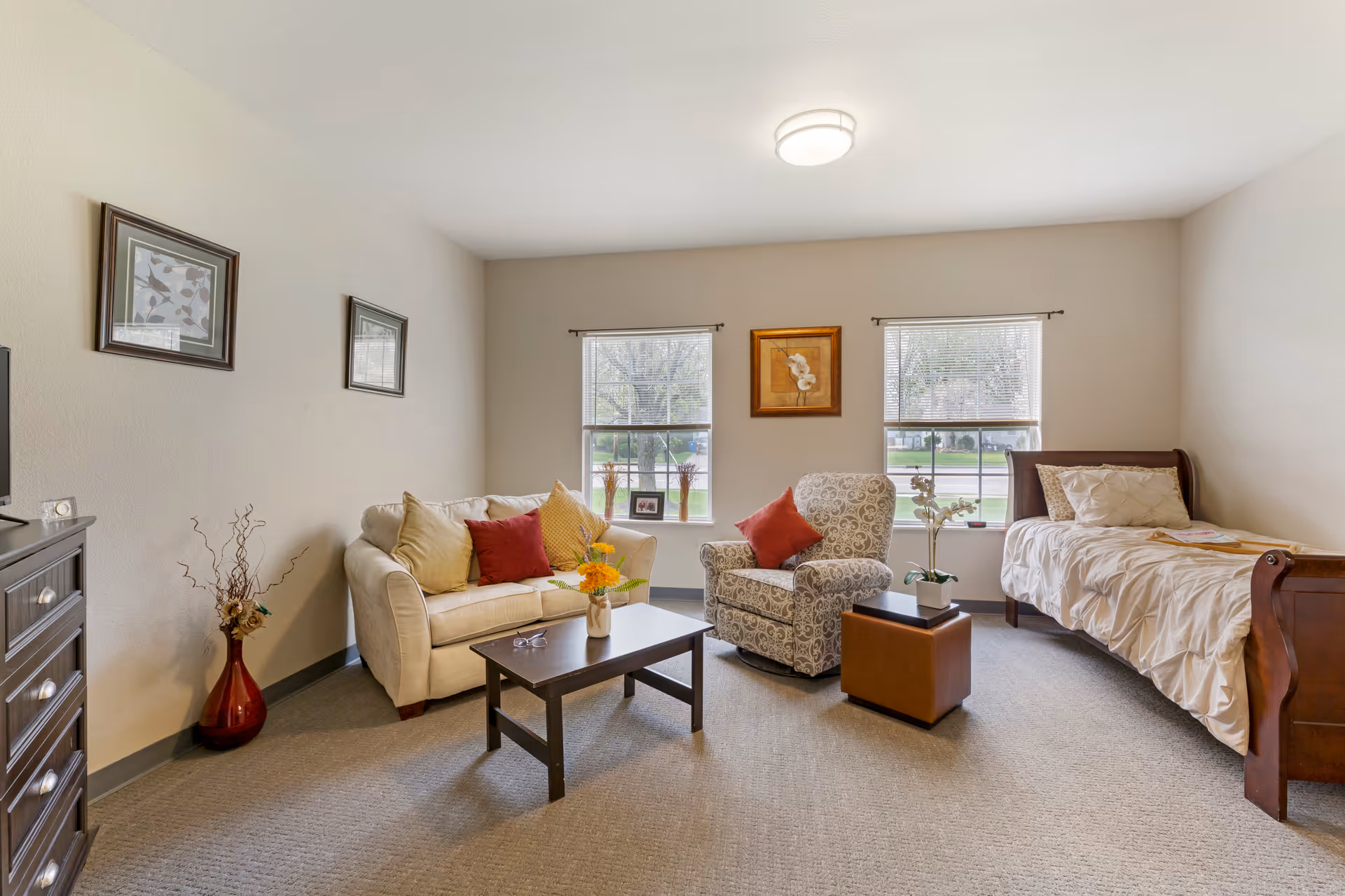 Bright furnished studio-style room with a sofa, armchair, coffee table, and a single bed by two windows.