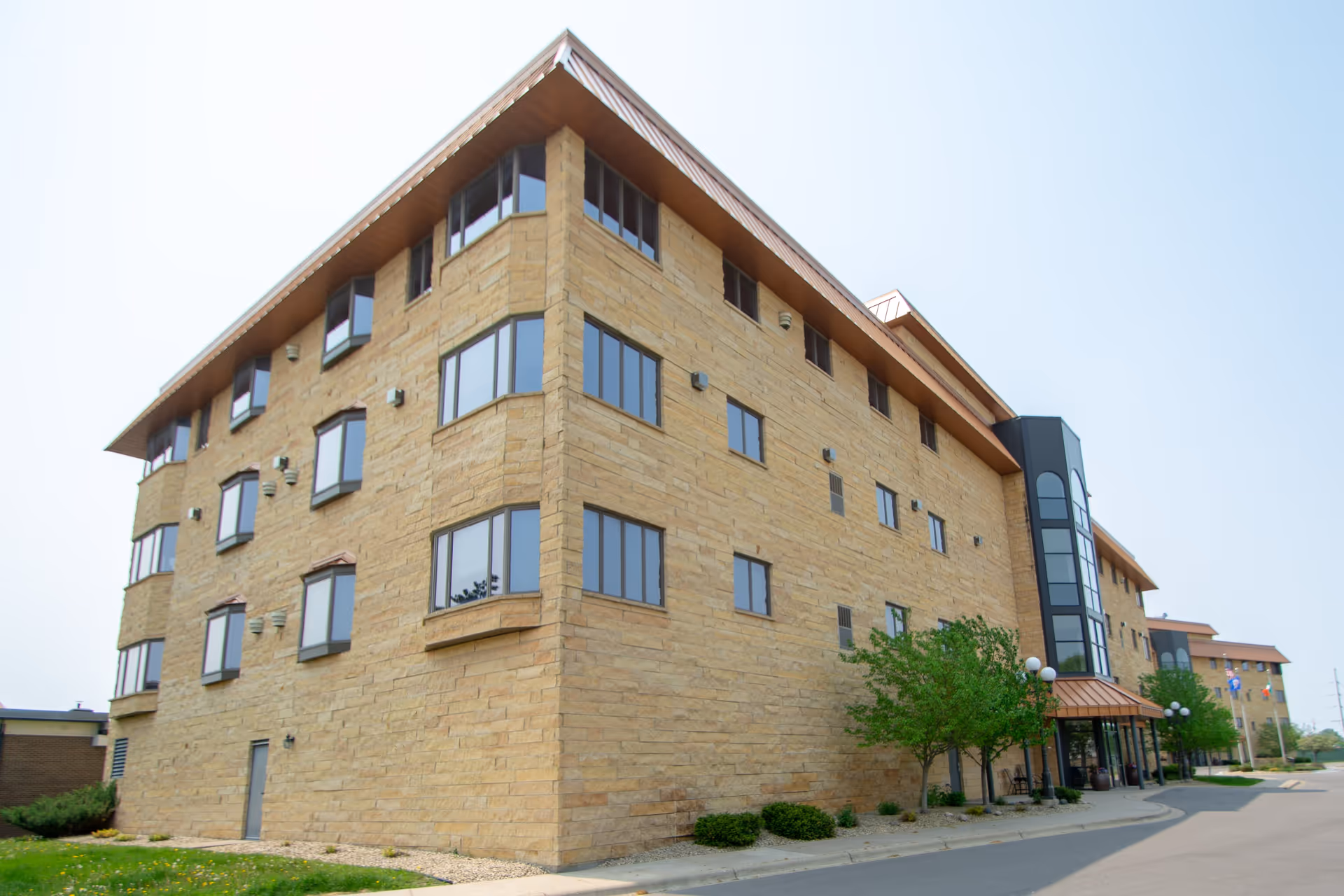 Exterior view of a multi-story senior living facility building with tan brick walls, multiple windows, and a glass entrance area. There are small trees and shrubs along the sidewalk in front of the building under a clear sky.