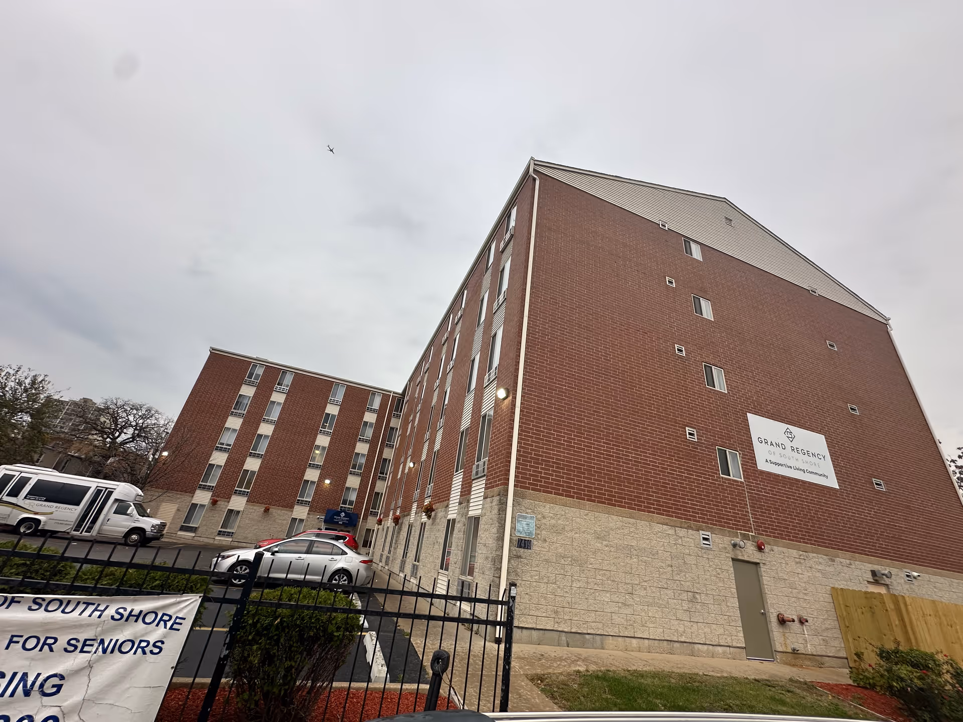 Exterior view of a multi-story brick and stone building with several windows, a parking lot with cars, and a sign that reads 'Grand Regency of South Shore A Supportive Living Community'. The sky is overcast.