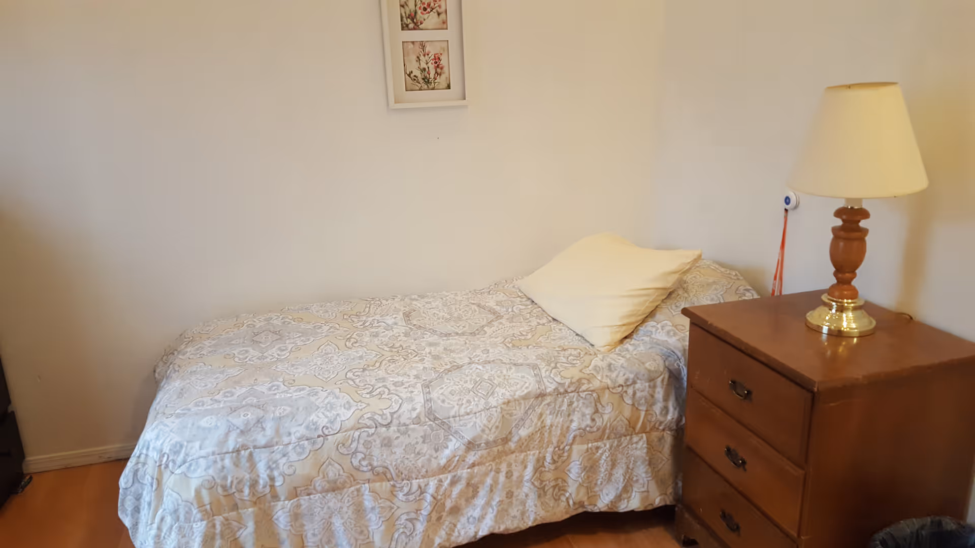 Small bedroom with a single bed covered in a patterned bedspread, a pillow, and a wooden nightstand with a lamp against a plain wall.