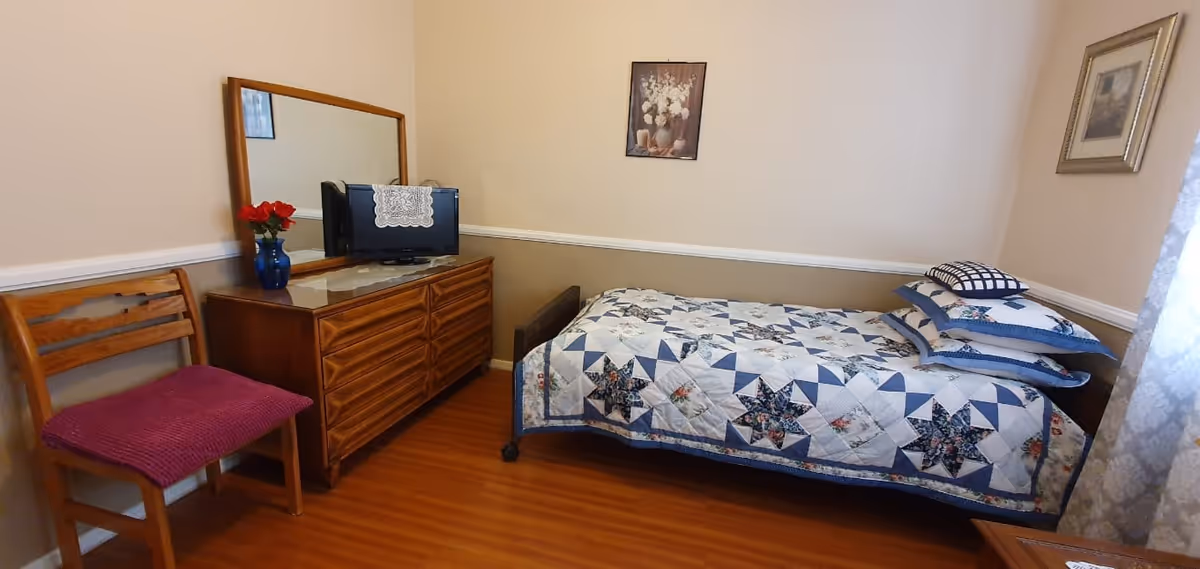 A small bedroom in a senior facility featuring a bed with quilt, dresser, chair, and TV.