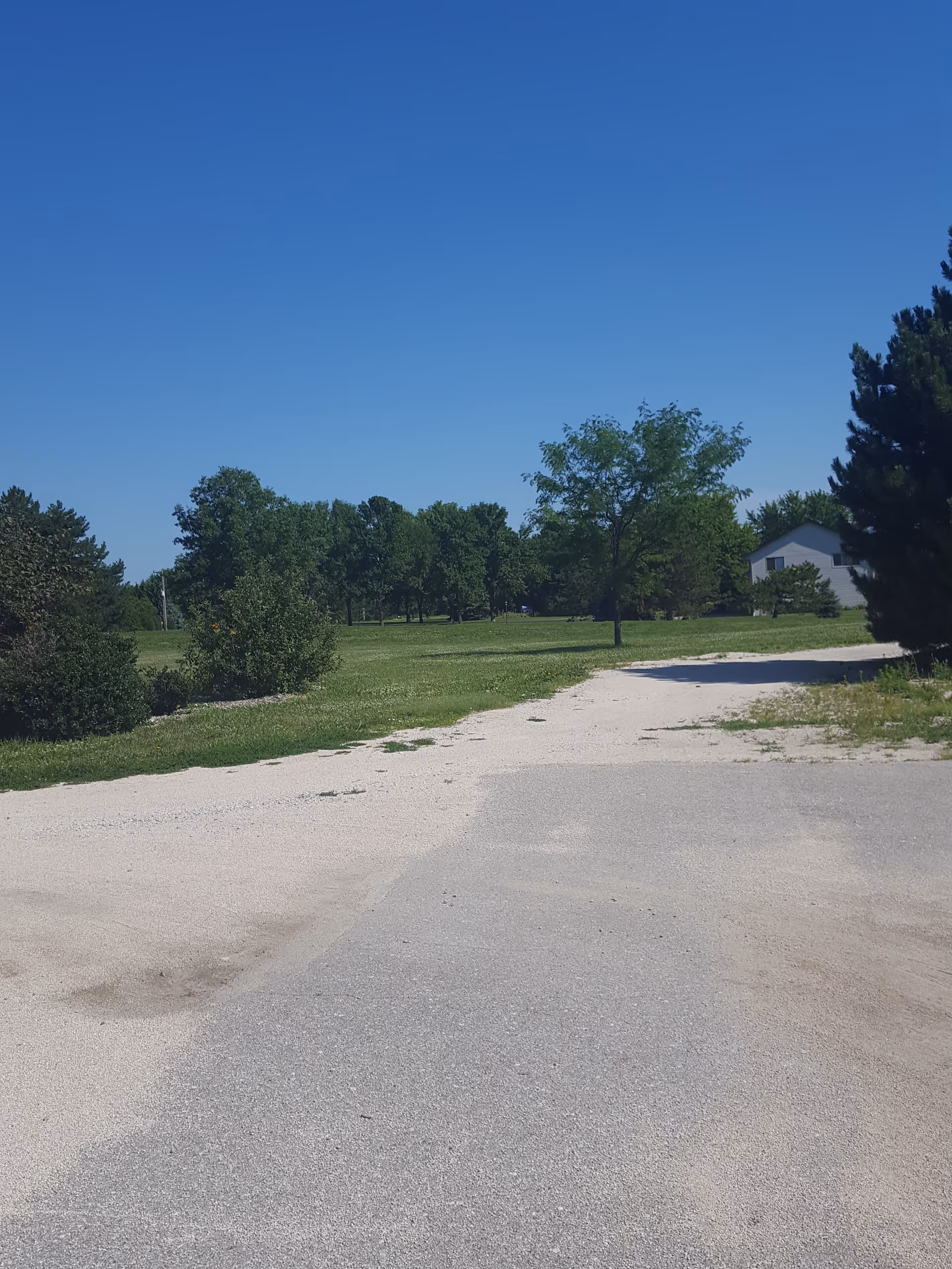 Gravel road leads to a grassy, tree-filled yard and a distant building.
