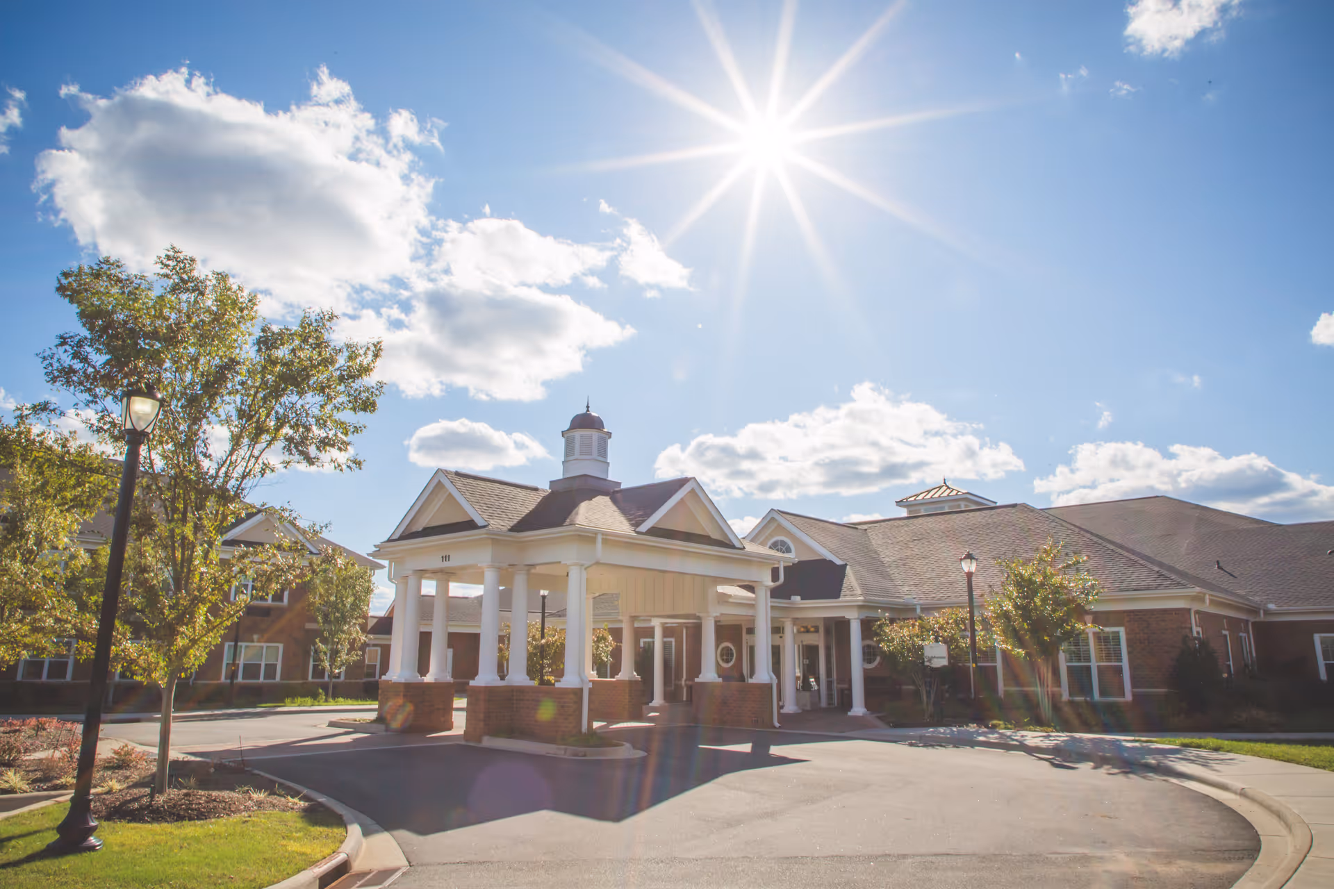 Exterior view of Wellmore of Tega Cay facility on a sunny day with a clear blue sky and scattered clouds. The building features a covered entrance with white columns and brick accents, surrounded by trees and a curved driveway.