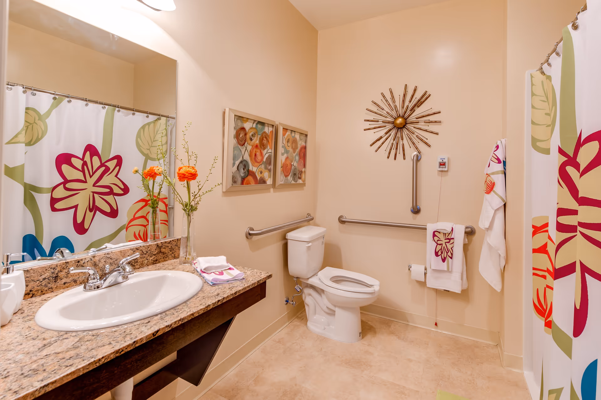 A clean and well-lit bathroom featuring a white sink with a granite countertop, a large mirror, a toilet with grab bars on the walls for accessibility, and a shower with a colorful floral curtain. The walls are decorated with two framed abstract floral paintings and a sunburst wall clock. Towels with floral designs hang near the toilet and shower.