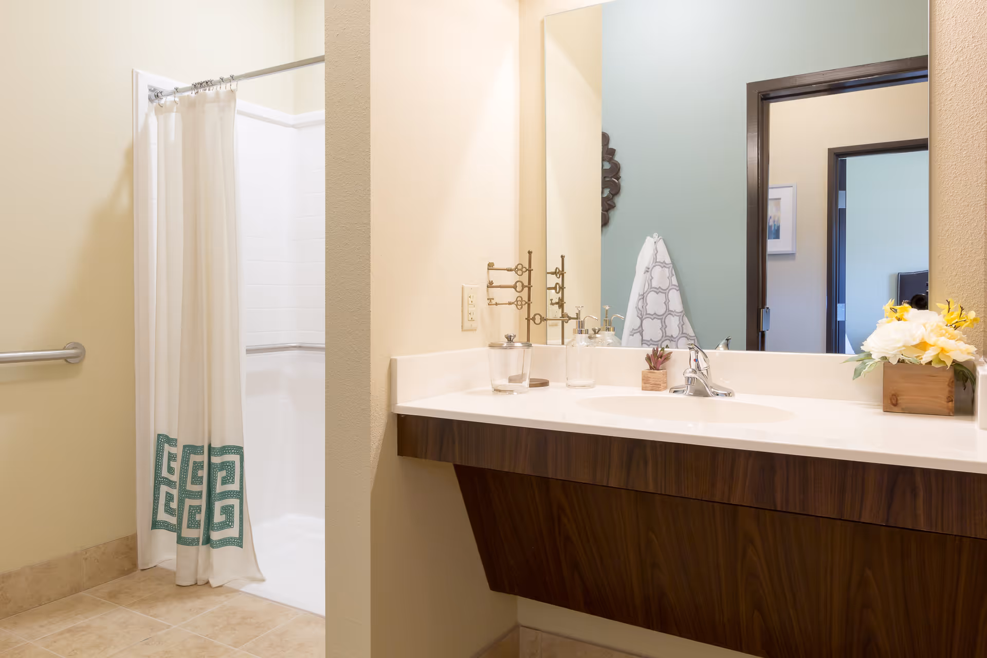 A bathroom with a white shower curtain featuring a green geometric pattern, a walk-in shower with white tiles, a wall-mounted grab bar, a wooden vanity with a white countertop, a sink with a faucet, a large mirror, a towel hanging on a hook, and a small flower arrangement on the counter.