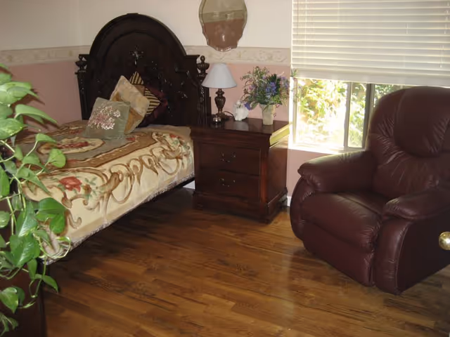 Bedroom with ornate dark wood bed, nightstand, lamp, plants, and a leather chair.