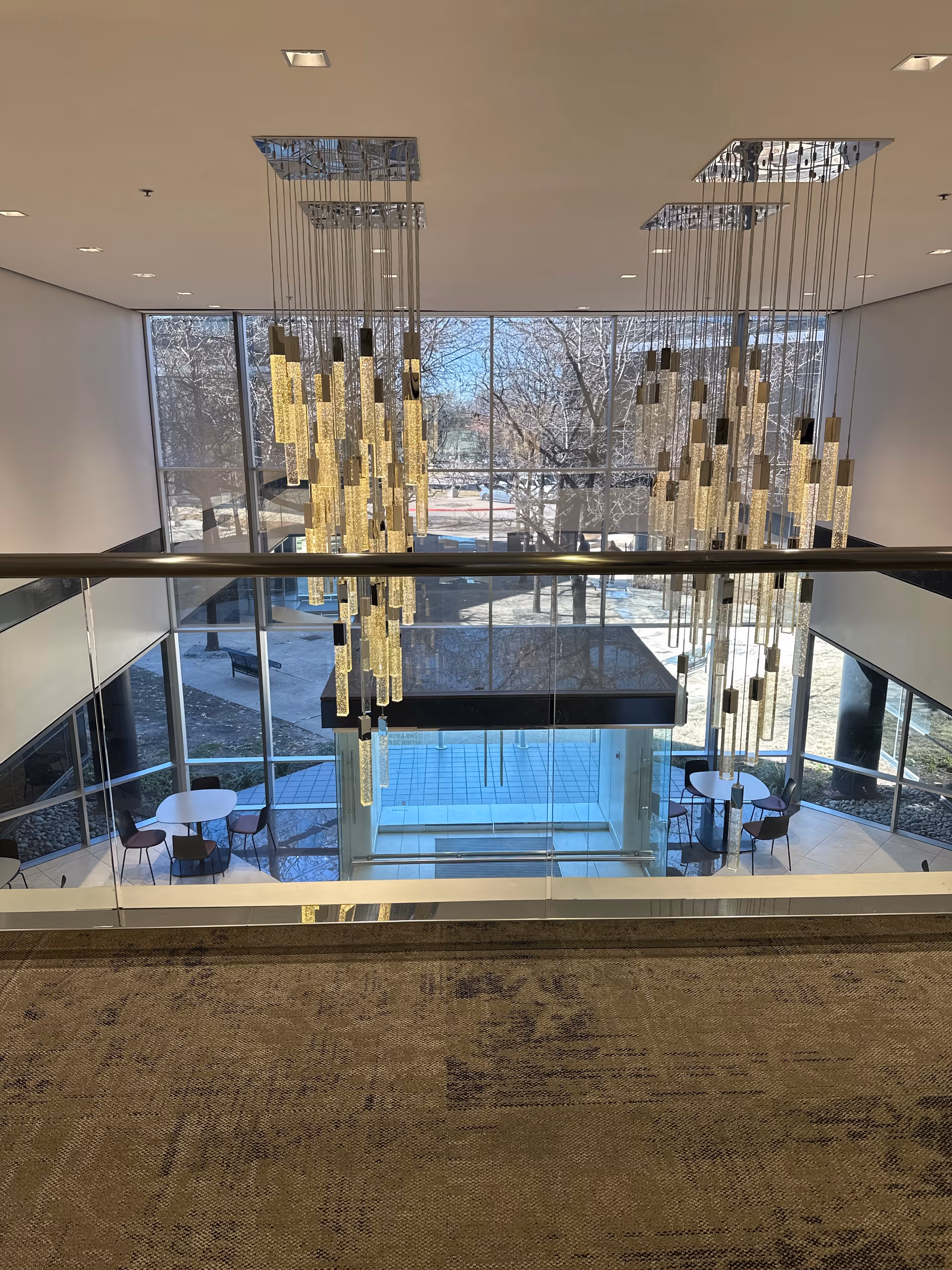Well-lit atrium lobby with glass railings, hanging chandeliers, and seating near large windows.