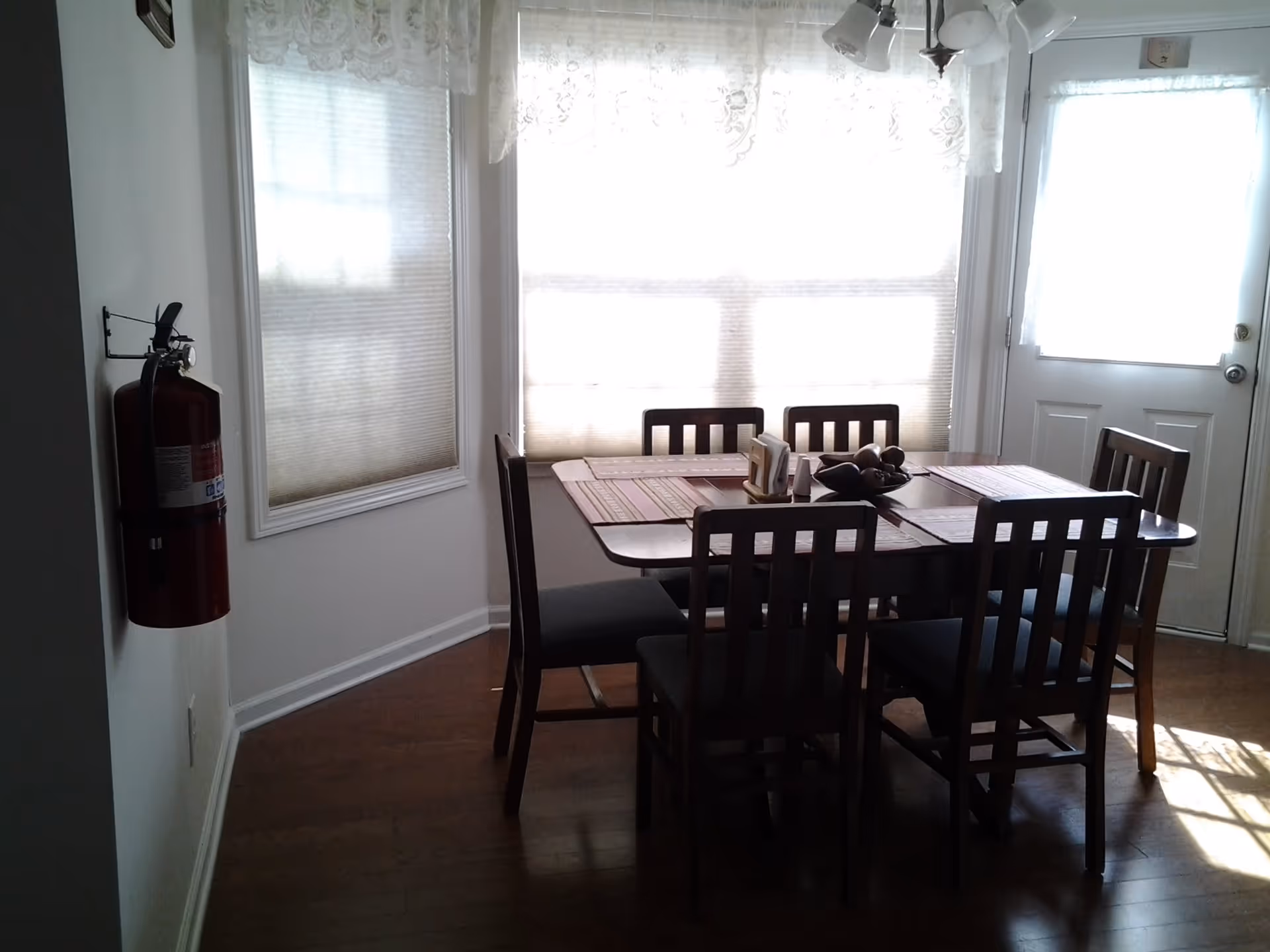 A dining area with a rectangular wooden table surrounded by six wooden chairs with dark cushions. The table is set with placemats, a napkin holder, salt and pepper shakers, and a decorative bowl with fruit. There are large windows with sheer white curtains and blinds letting in natural light. A fire extinguisher is mounted on the wall to the left, and a door with a window is visible on the right side.