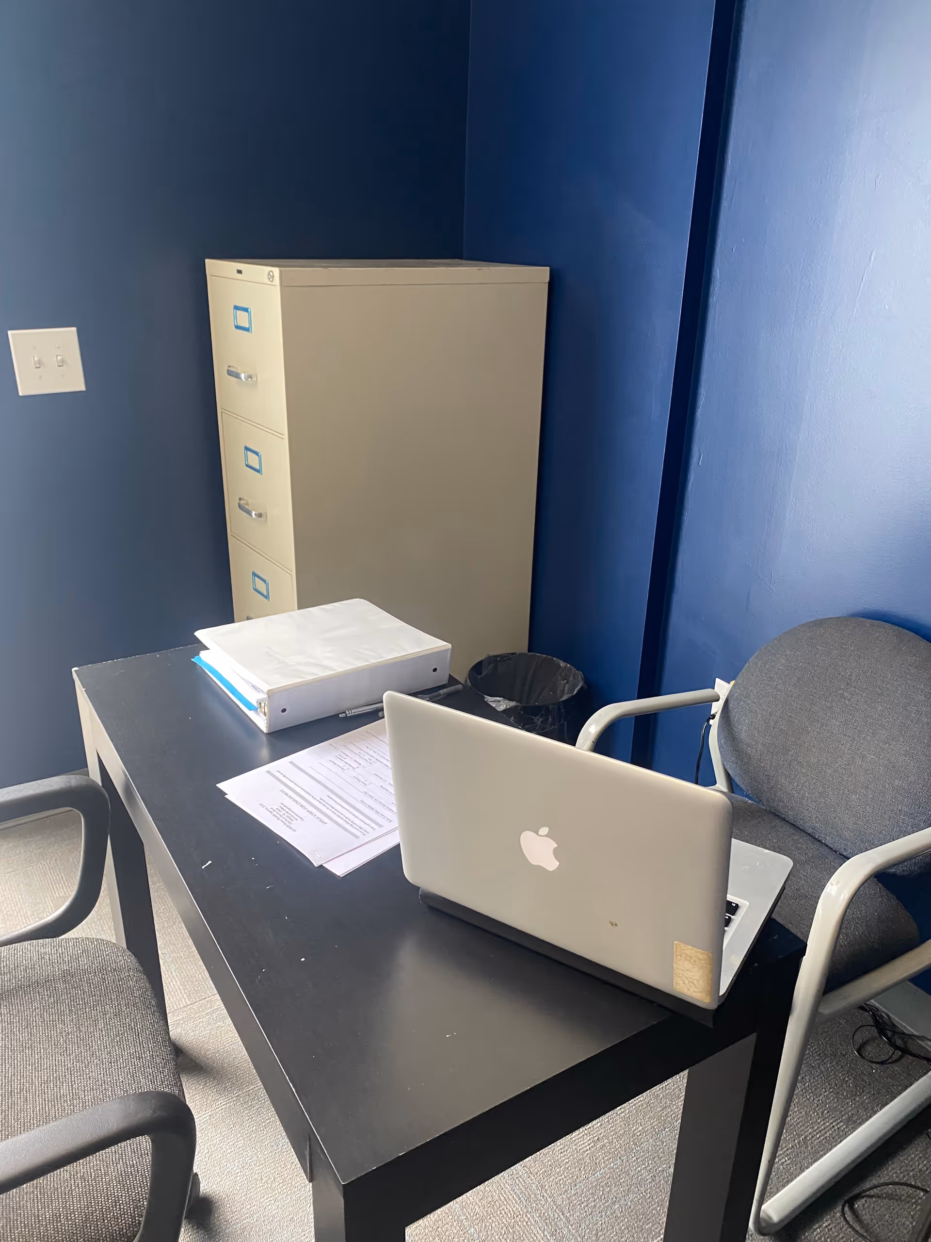 Office workspace with blue walls, desk, laptop, filing cabinet, and two chairs.