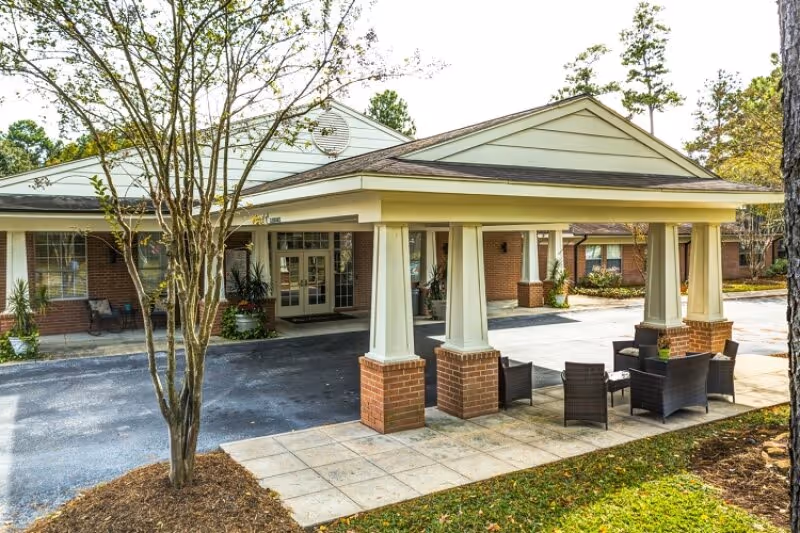 Exterior view of Cypresswood Memory Care facility showing a covered entrance with four large columns on brick bases, outdoor seating with chairs and a table on a paved area, and surrounding trees and landscaping.