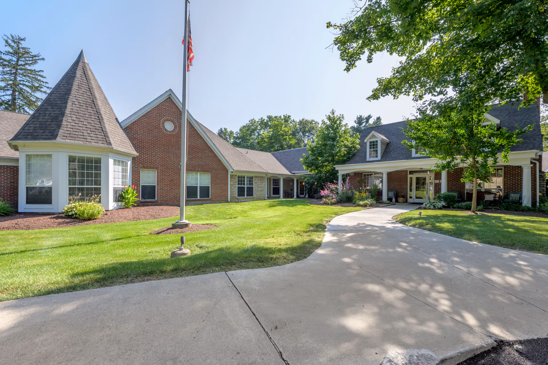 Exterior view of a senior living facility building with a brick facade, a peaked roof, and a covered entrance. There is a concrete walkway leading to the entrance, a well-maintained lawn, trees, and an American flag on a flagpole in front of the building under a clear blue sky.